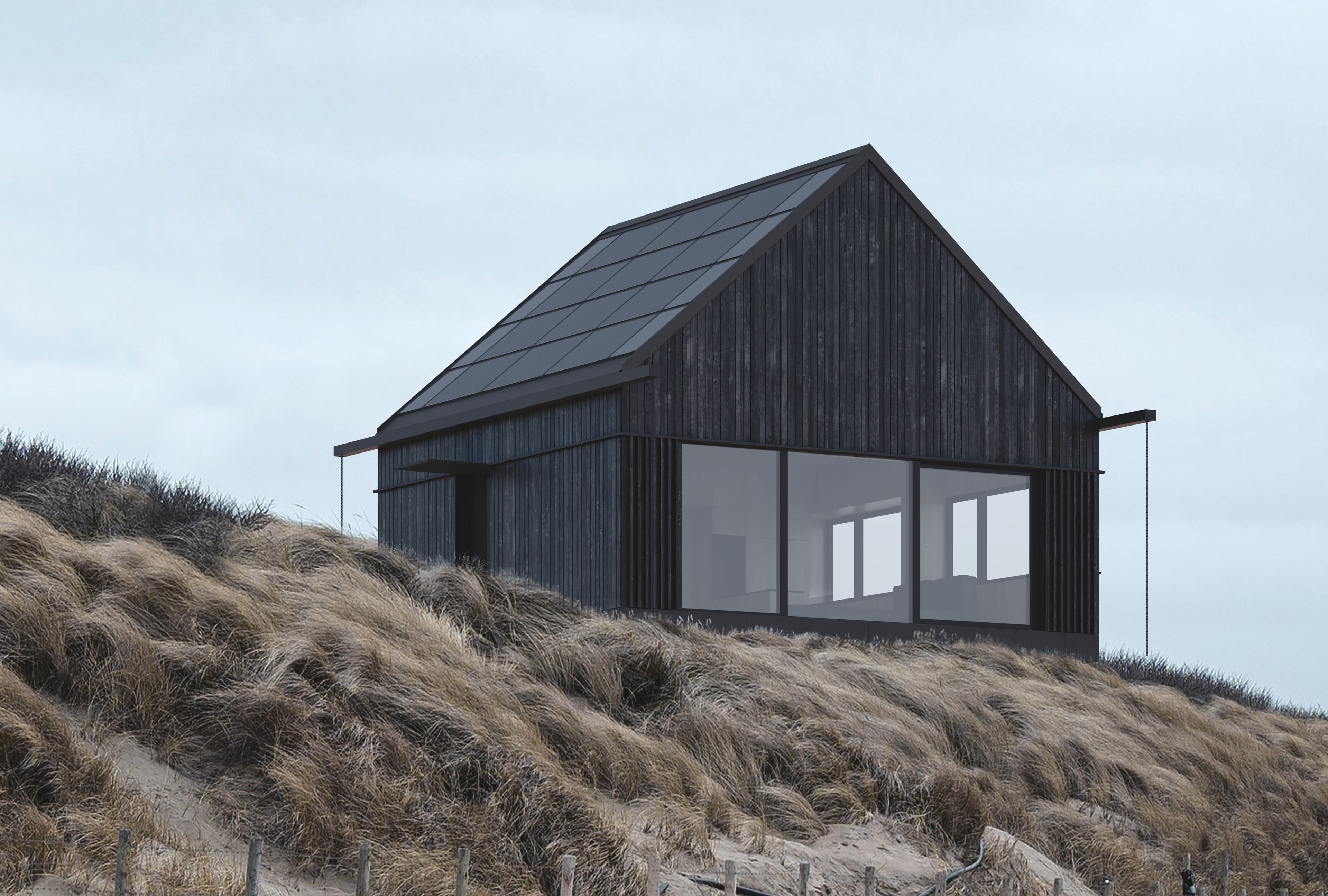 A modern, black house with a solar roof stands on a hill covered with tall, dry grasses under a cloudy sky.