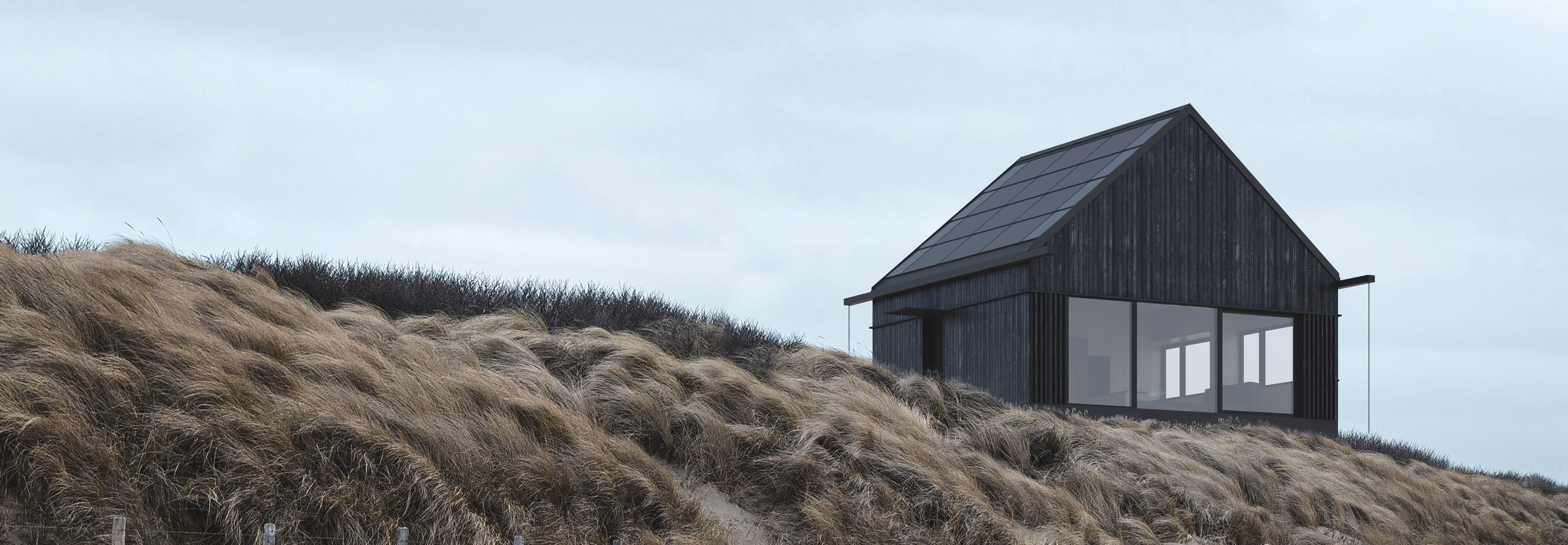 A modern, black house with a solar roof stands on a hill covered with tall, dry grasses under a cloudy sky.