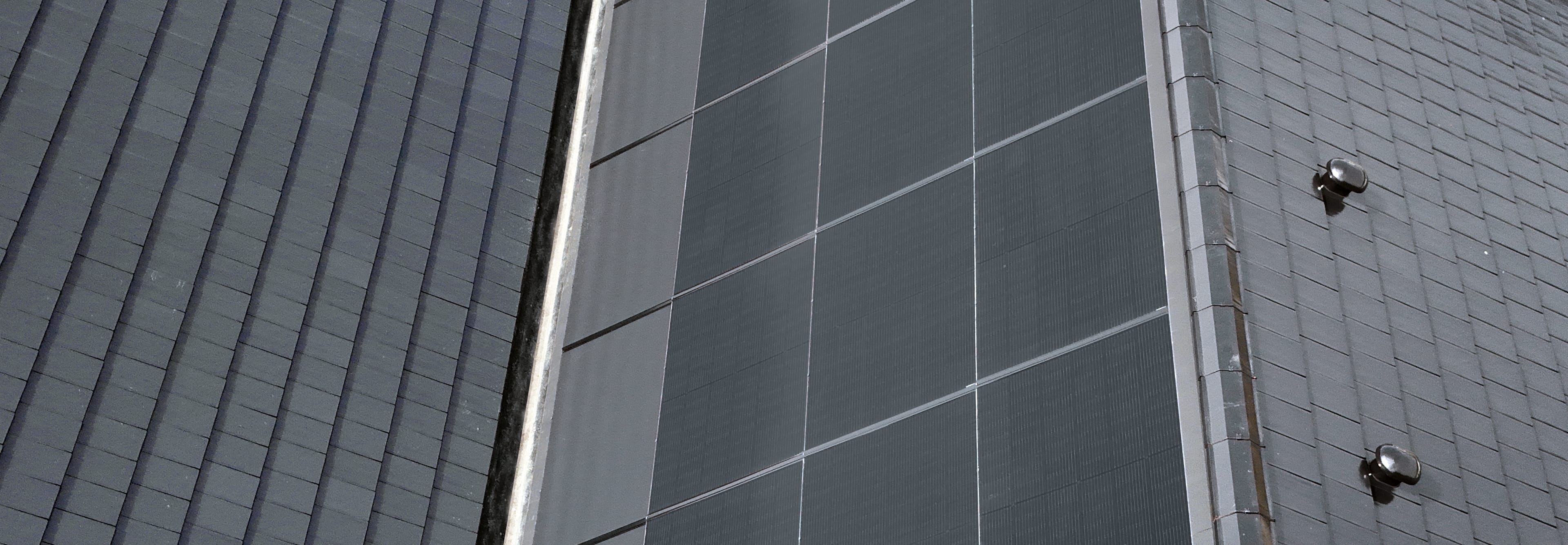 Zbliżenie na połacie dachu, pokazujące połączenie ciemnych, gładkich paneli fotowoltaicznych z teksturowanymi, szarymi dachówkami.