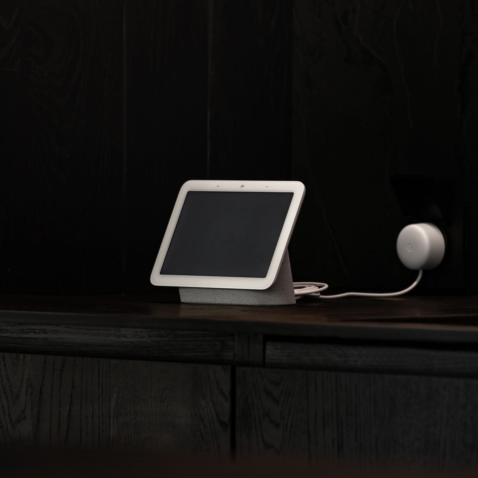 A white smart display with a blank screen stands on a dark shelf, connected to a round power adapter in an outlet.