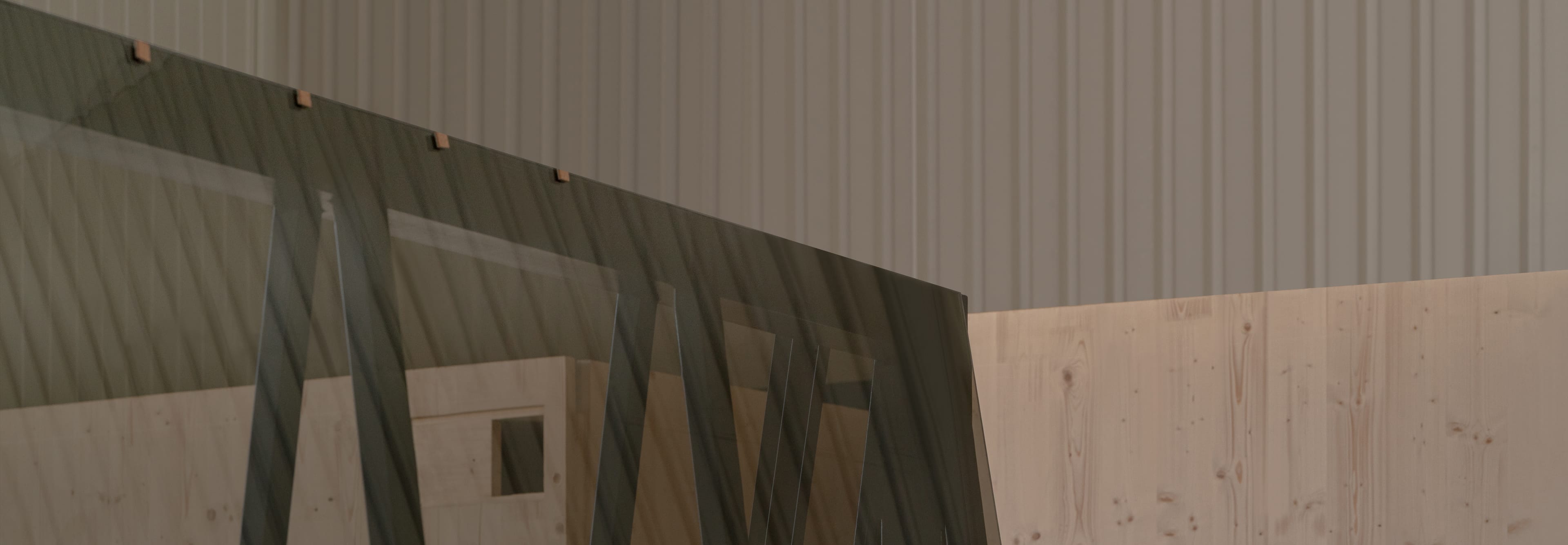Large sheets of glass stand against a wall of light-colored construction wood in a hall. Above, a wall of corrugated iron is visible.