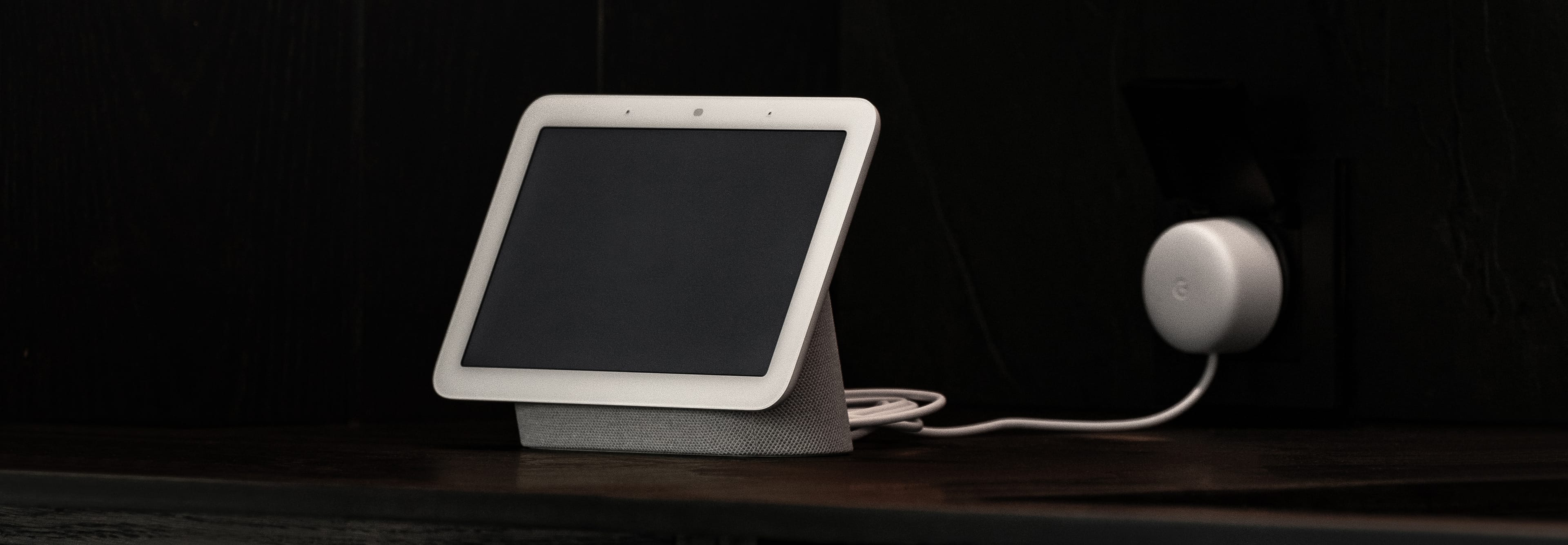 A white smart display with a blank screen stands on a dark shelf, connected to a round power adapter in an outlet.