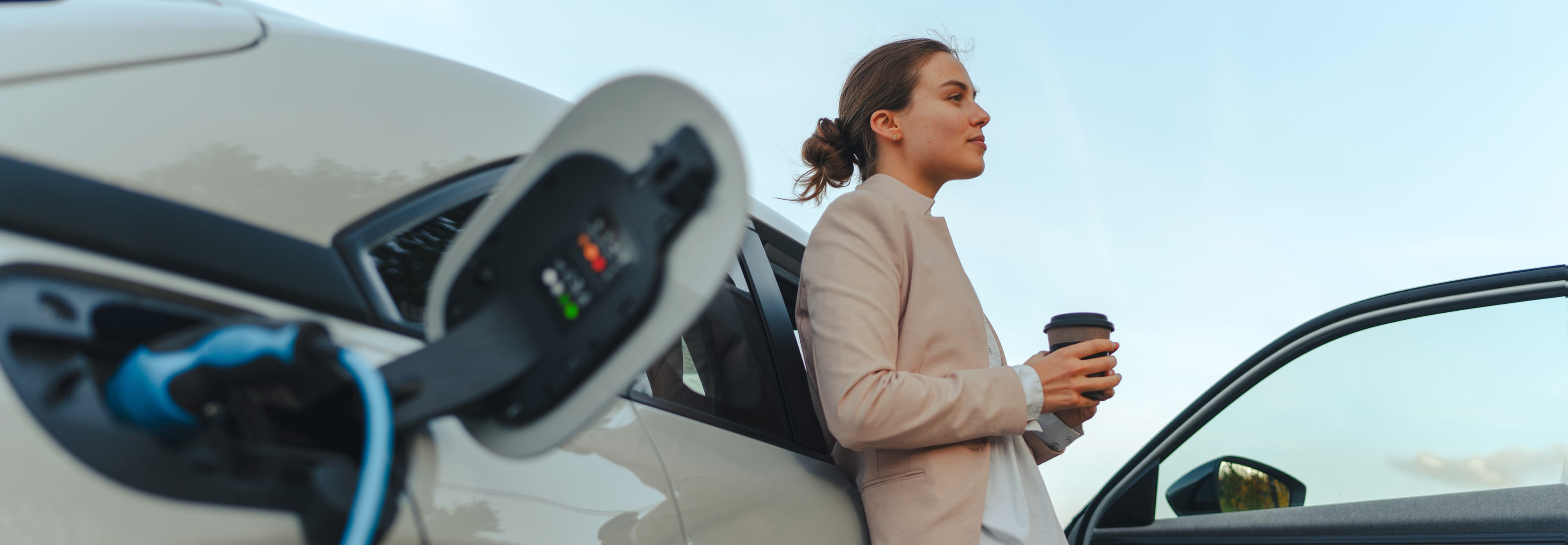 A woman with coffee is leaning against a white electric car that is being charged. In the foreground, a charging port with a plug is visible.