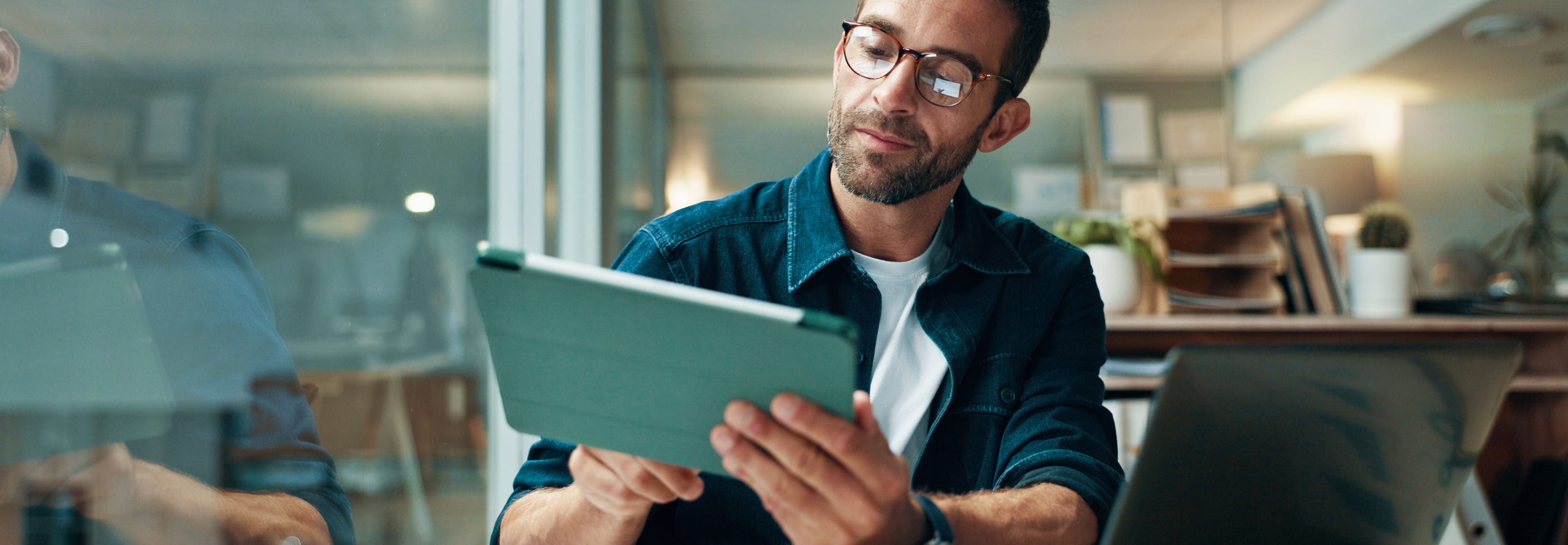A man with glasses and a blue shirt sits in an office, holding a tablet. An open laptop is on the desk next to him.