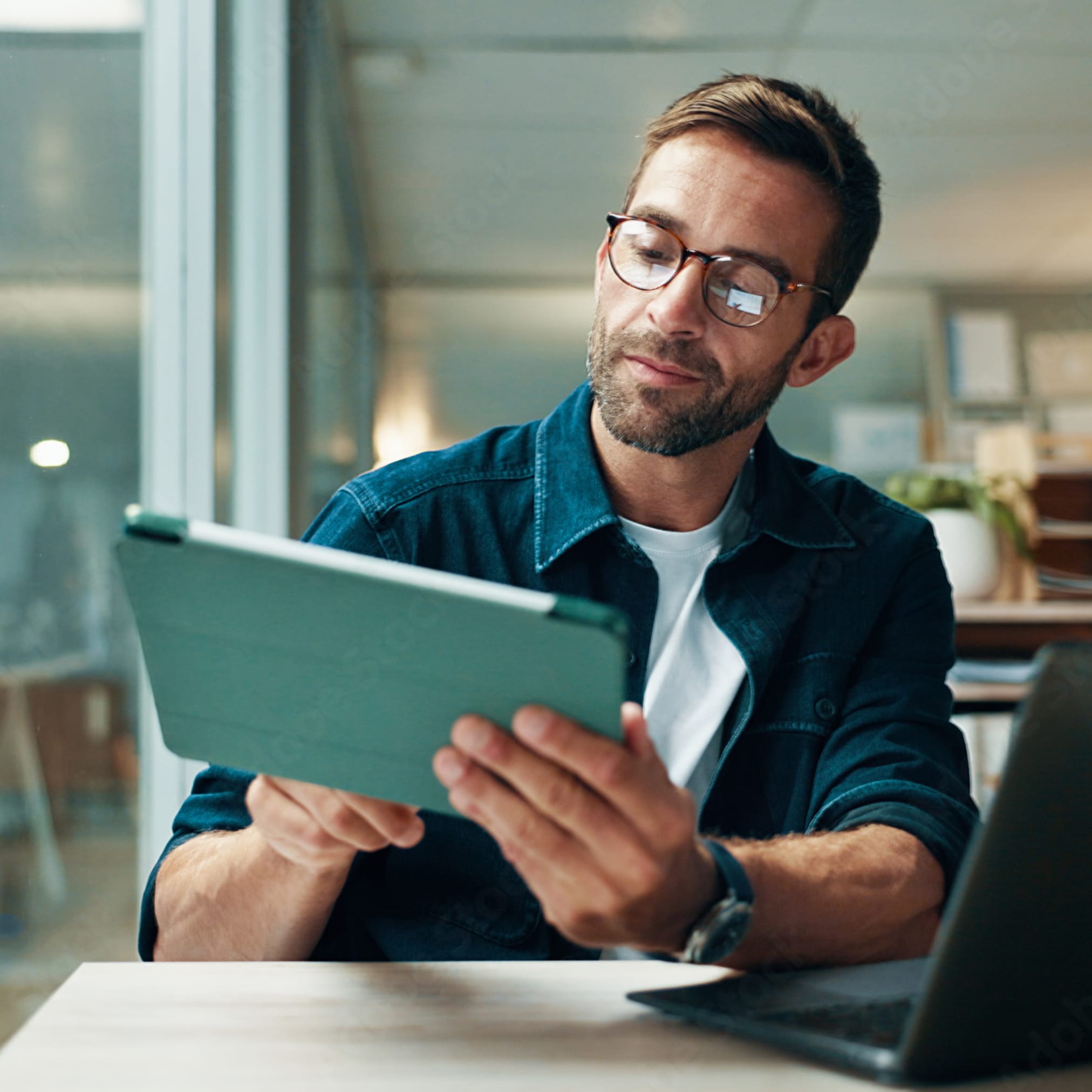 A man with glasses and a blue shirt sits in an office, holding a tablet. An open laptop is on the desk next to him.