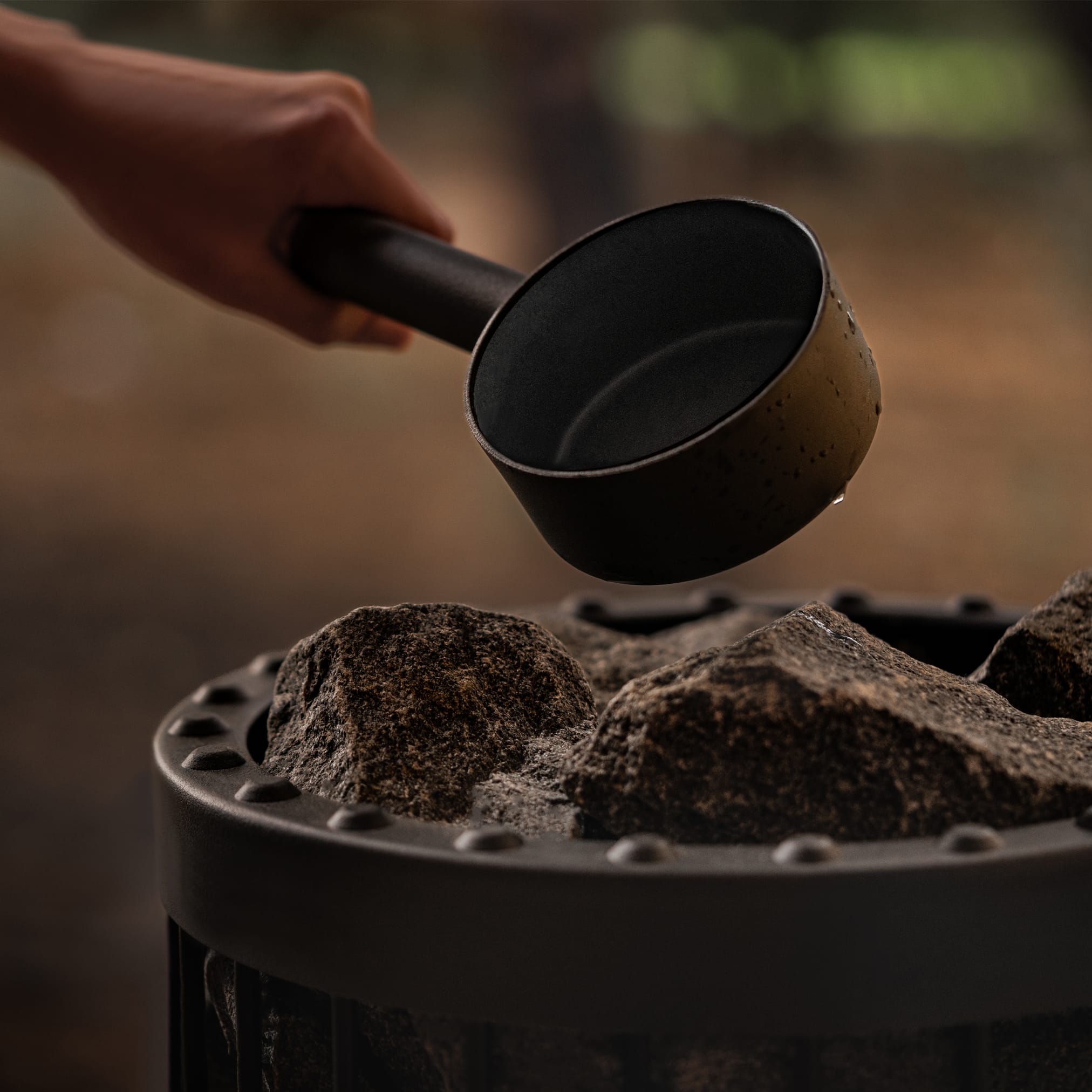 Close-up of a hand holding a black ladle from which water drips onto hot stones in a sauna stove.