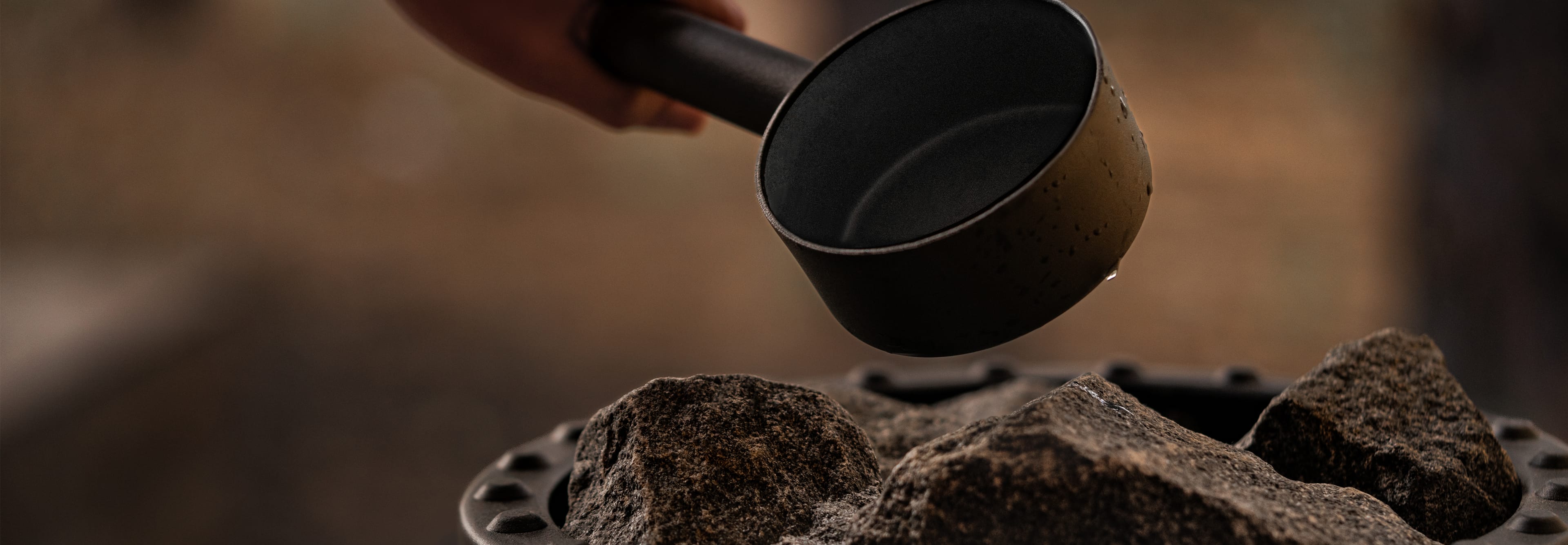 Close-up of a hand holding a black ladle from which water drips onto hot stones in a sauna stove.