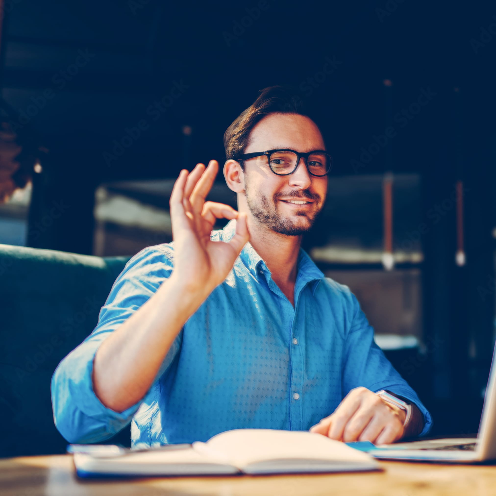 A smiling man with glasses and a blue shirt is sitting at a laptop, showing an "OK" gesture with his right hand.