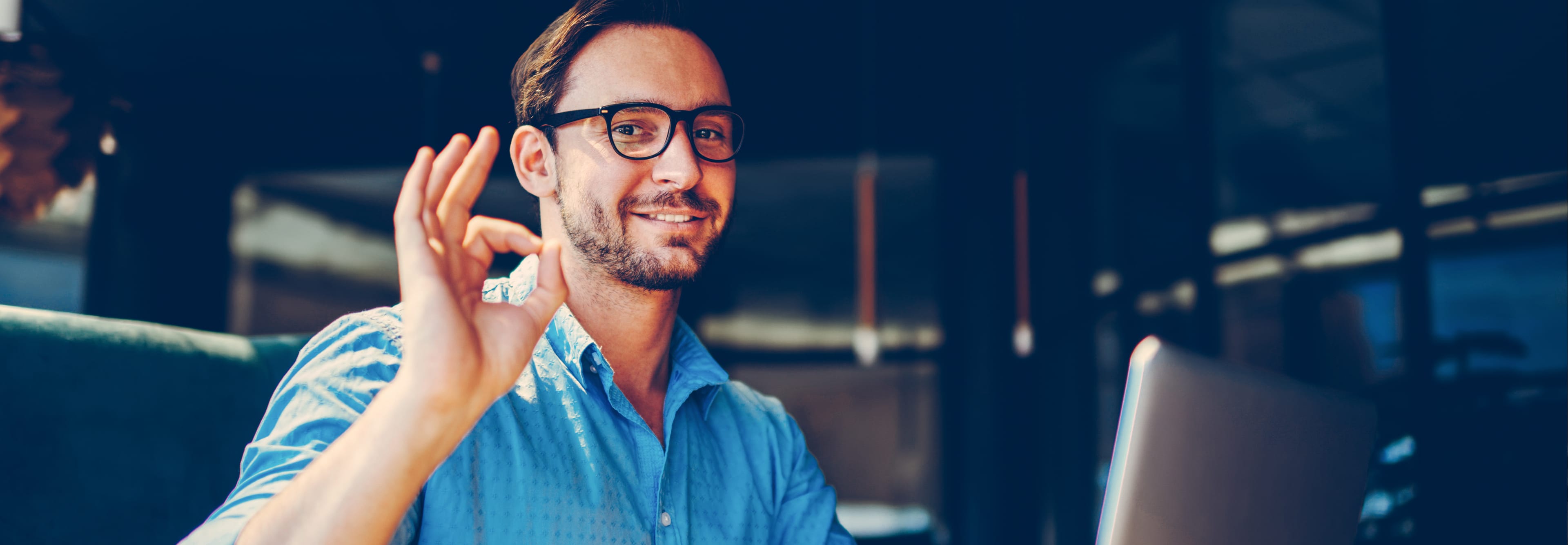 A smiling man with glasses and a blue shirt is sitting at a laptop, showing an "OK" gesture with his right hand.