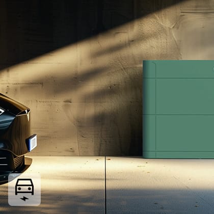 The front of a dark electric car pulls up to a green energy storage unit in a sunlit garage.