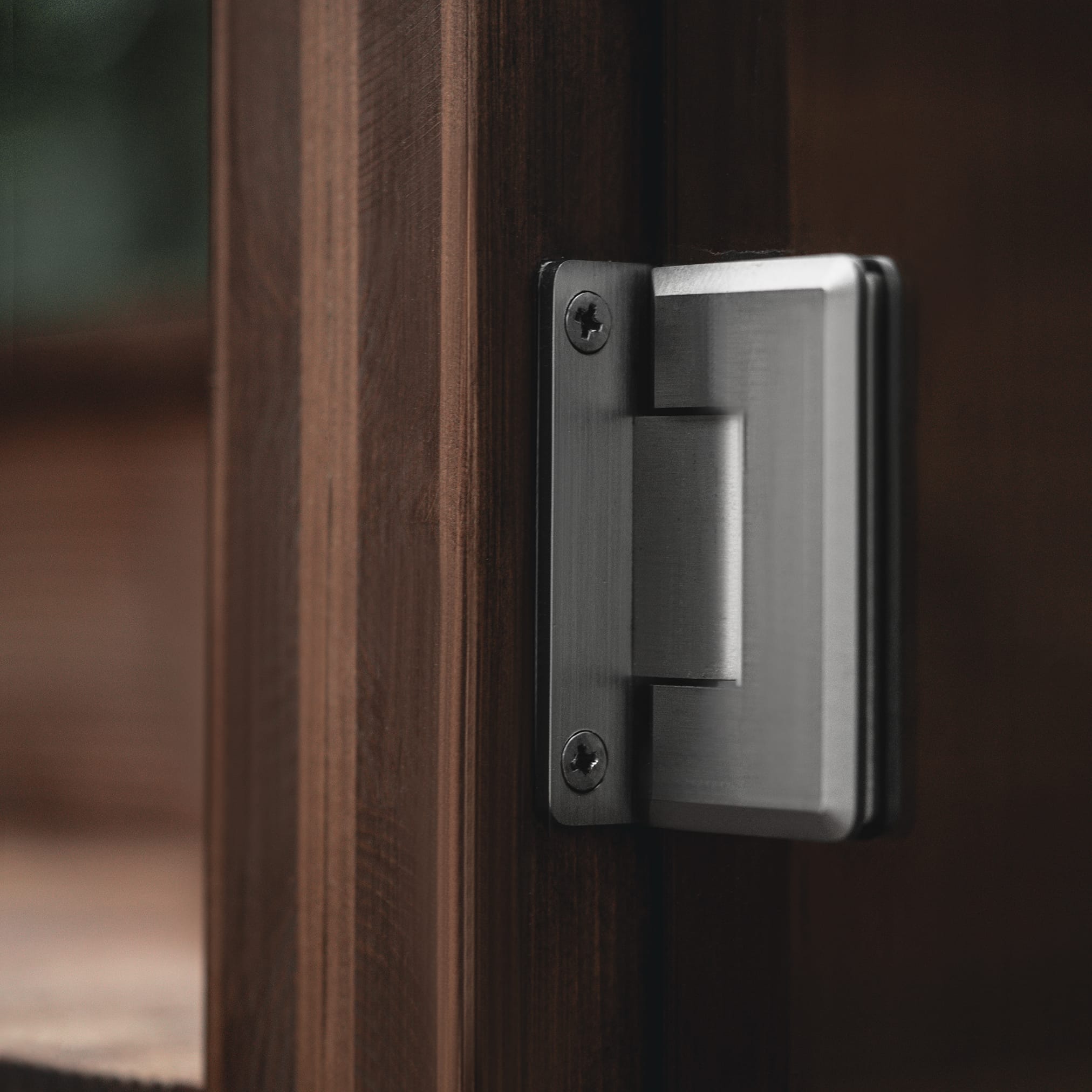 Close-up of a massive, graphite-colored hinge made of brushed metal, attached with two screws to a dark, brown wood frame.