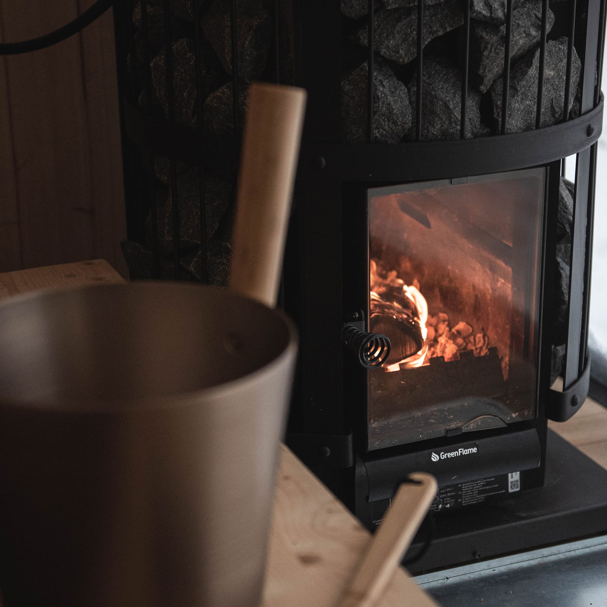 A modern, black sauna stove with a glass door, behind which a fire is burning. Stones are on the stove, and a sauna bucket is in the foreground.
