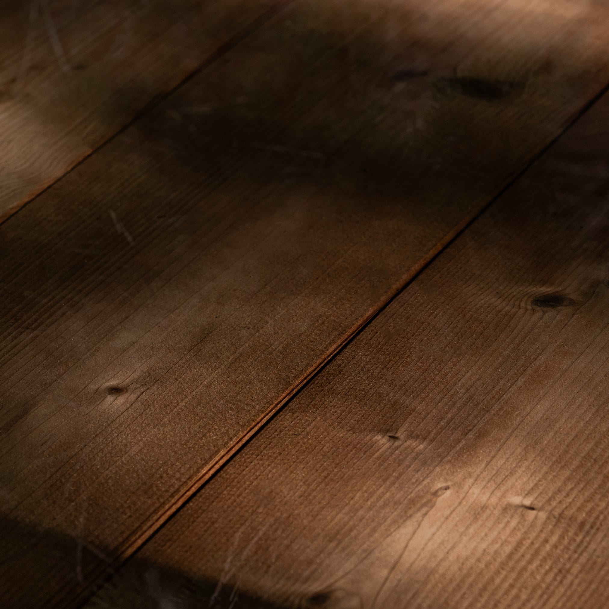 Warm, brown floorboards photographed up close at an angle. Visible grain, knots, and light and shadow falling on them diagonally.