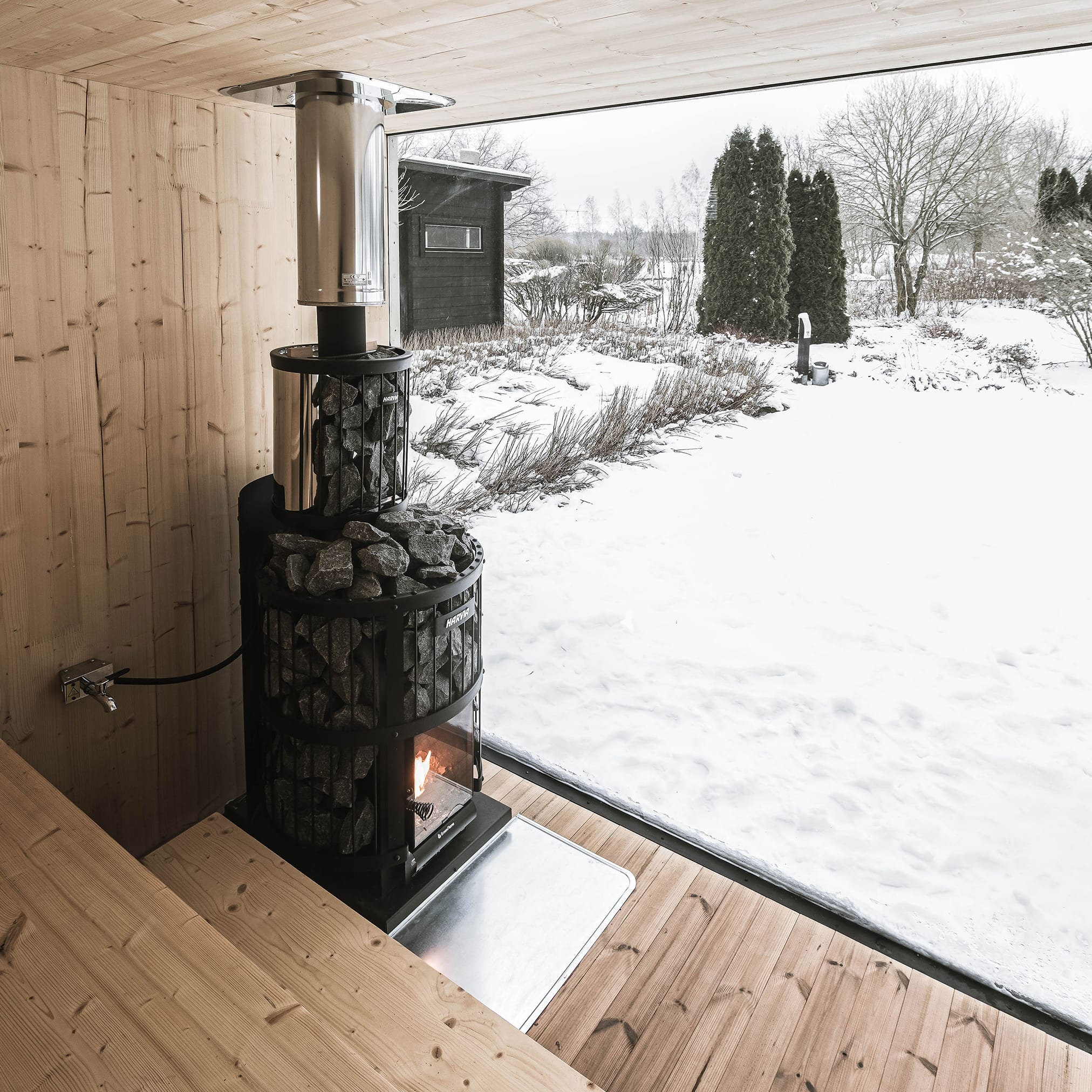 The interior of a wood-paneled sauna with a black, cylindrical stone stove in which a fire is visible. A large, panoramic window shows a snowy, winter garden.