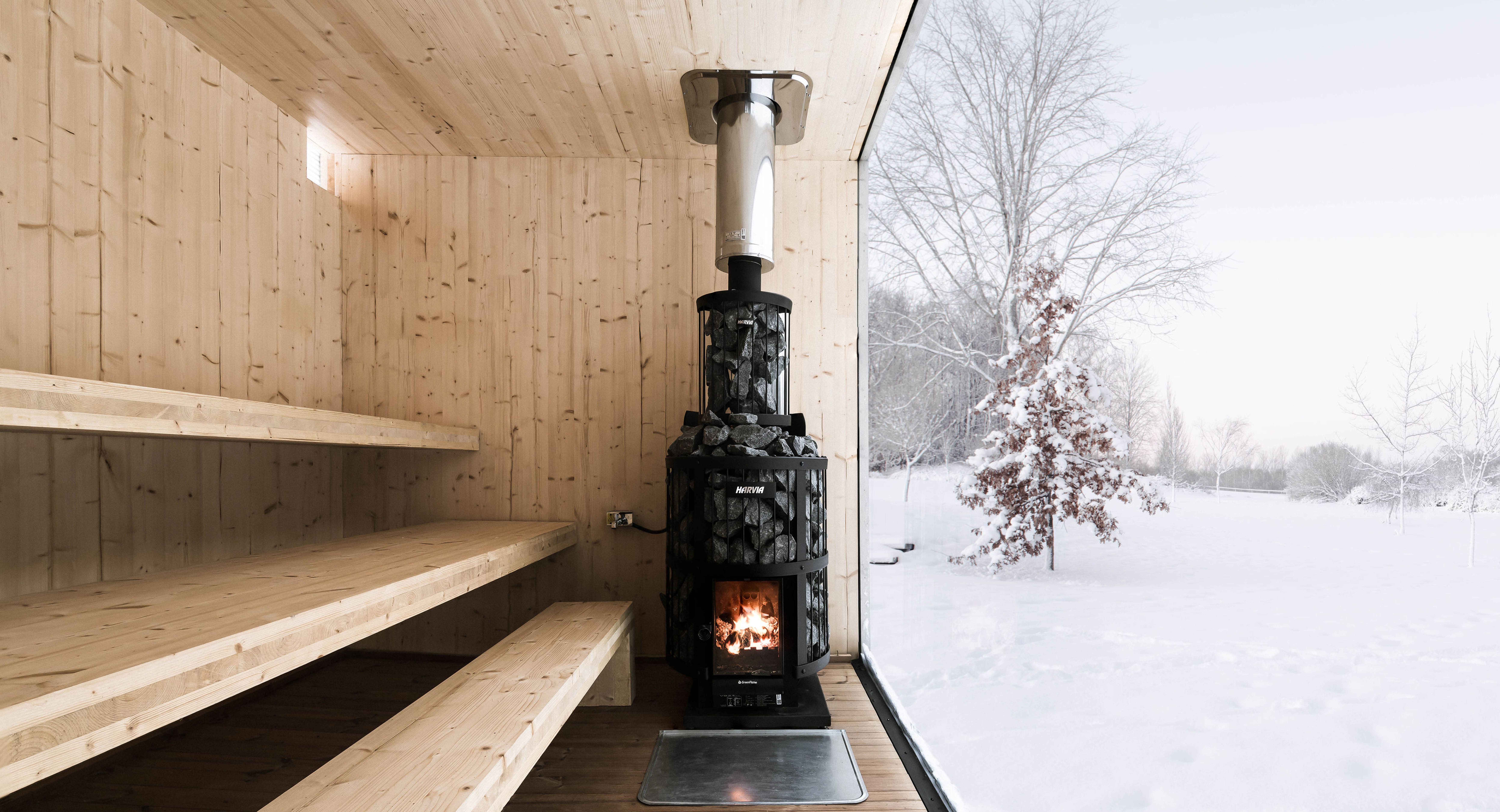 The interior of a sauna finished in light wood, with a black wood-burning stove and a large window with a view of a snowy landscape.