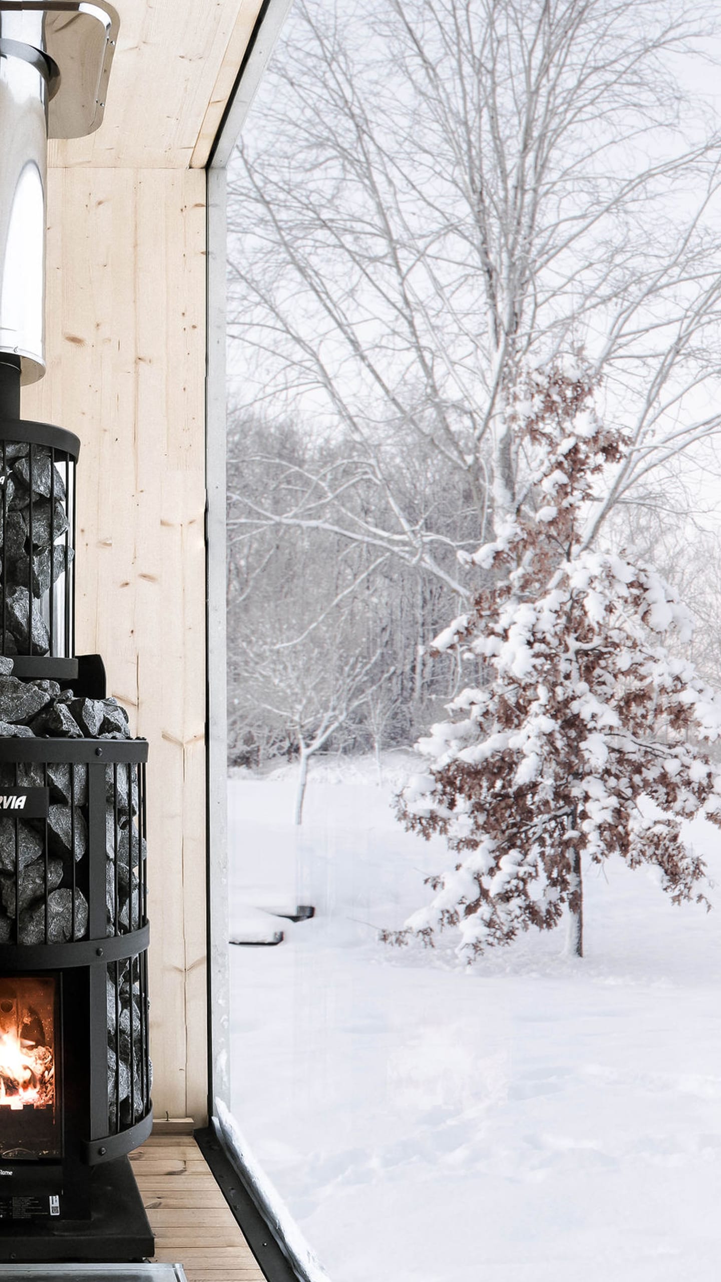 The interior of a sauna finished in light wood, with a black wood-burning stove and a large window with a view of a snowy landscape.