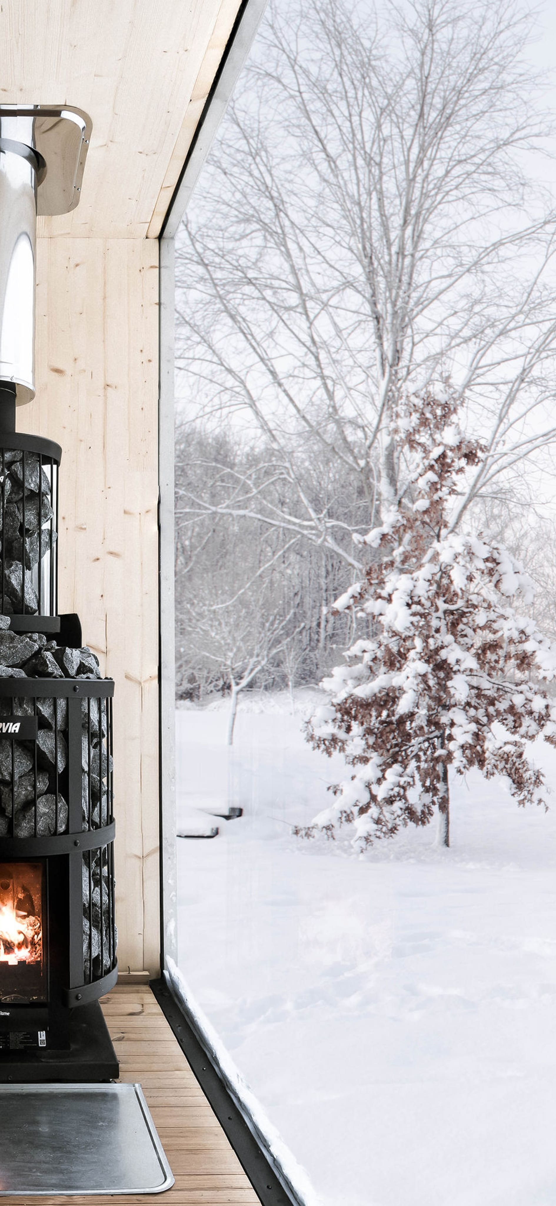 The interior of a sauna finished in light wood, with a black wood-burning stove and a large window with a view of a snowy landscape.