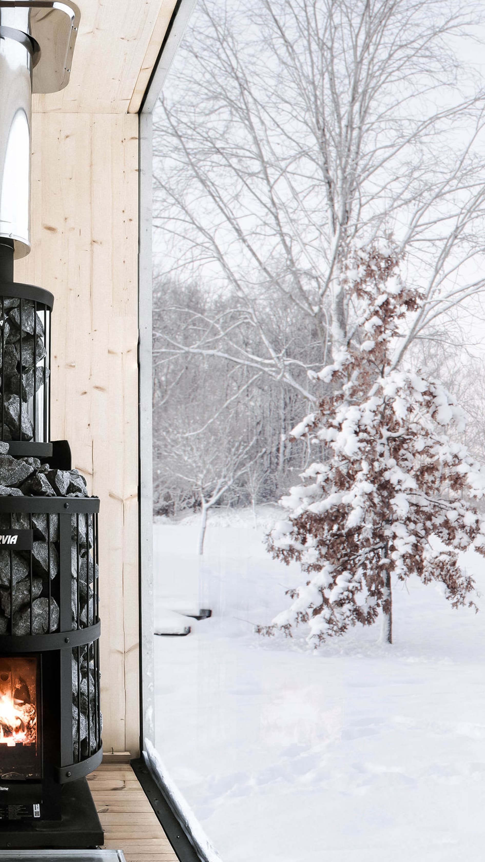 The interior of a sauna finished in light wood, with a black wood-burning stove and a large window with a view of a snowy landscape.