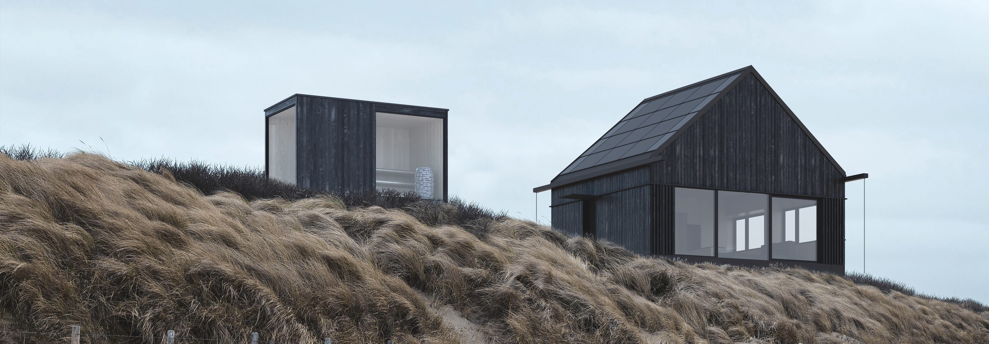 Dwa budynki- dom i sauna z czarnego drewna na wzgórzu porośniętym suchą trawą. Jeden ma spadzisty dach, a drugi ma kształt sześcianu.