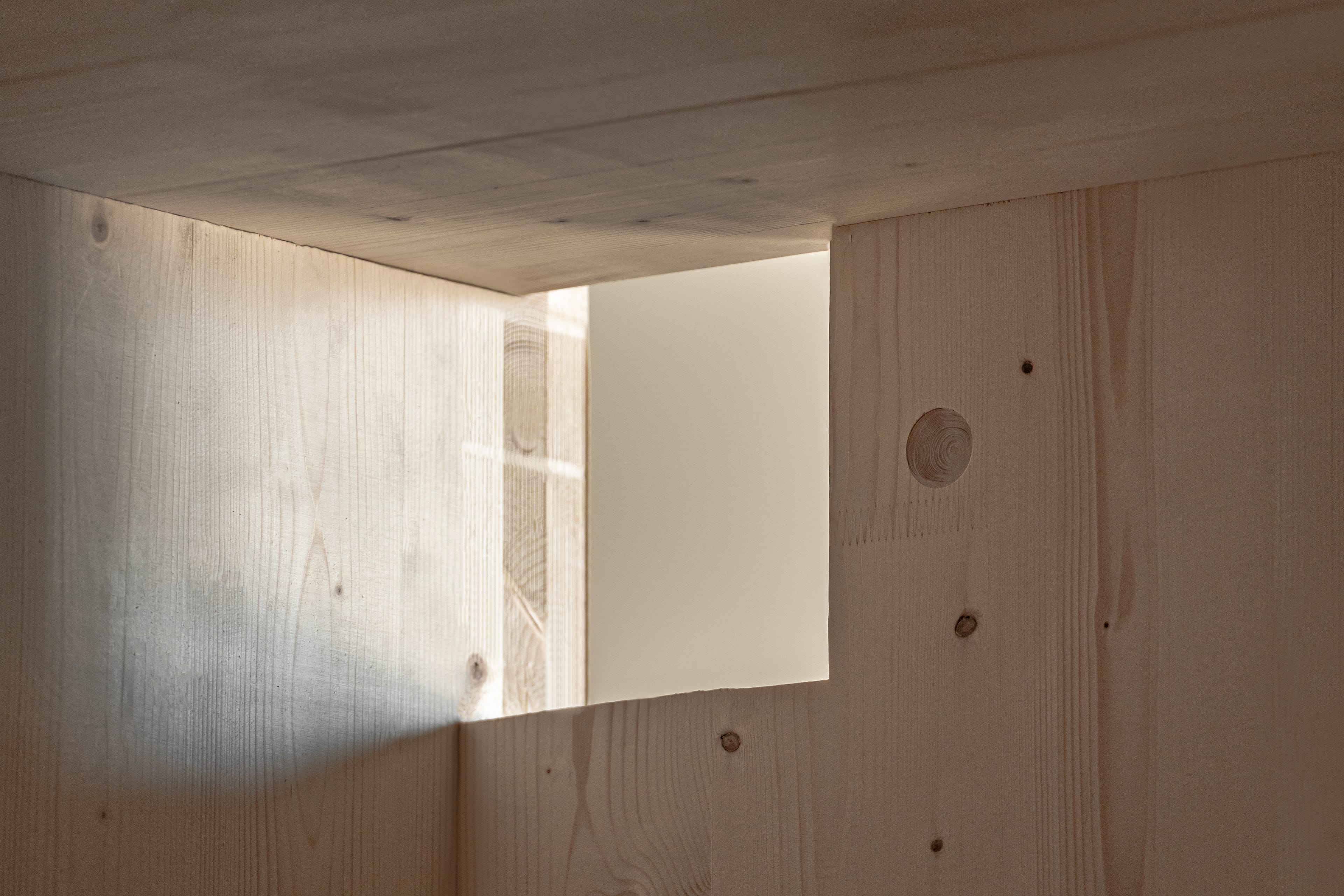 Close-up of a corner of a light wood structure. Light enters through a rectangular opening, creating a bright spot on the wooden wall.