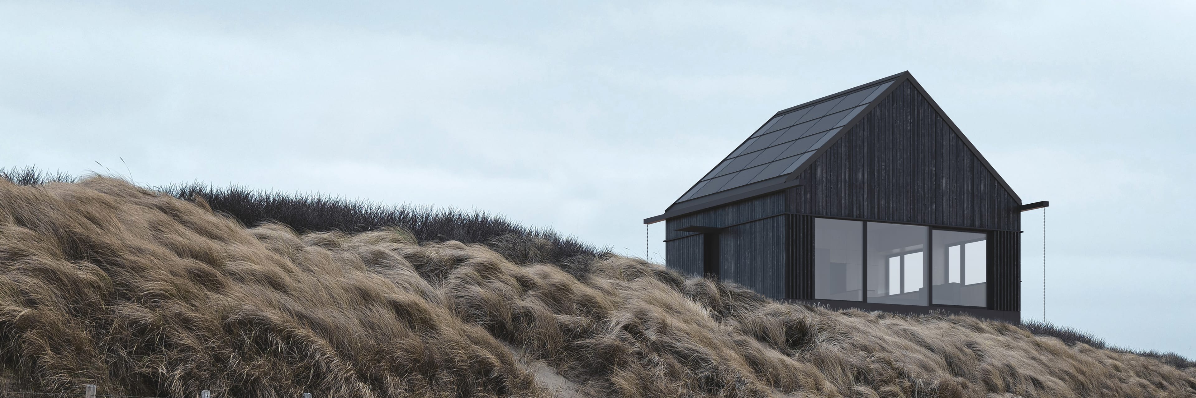 A dark, modern house with a roof entirely covered with photovoltaic panels, standing on a meadow at the foot of forested mountains.