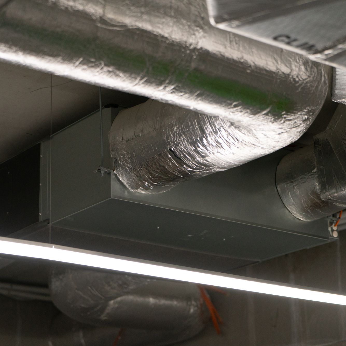 Thick, round ducts made of silvery material enter a steel box suspended from the ceiling. In the background, you can see the ducts coming out of the steel box.