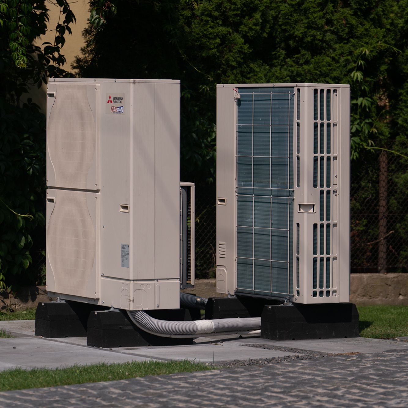 Two heat pumps standing next to each other, placed on concrete slabs. Greenery can be seen in the surroundings.