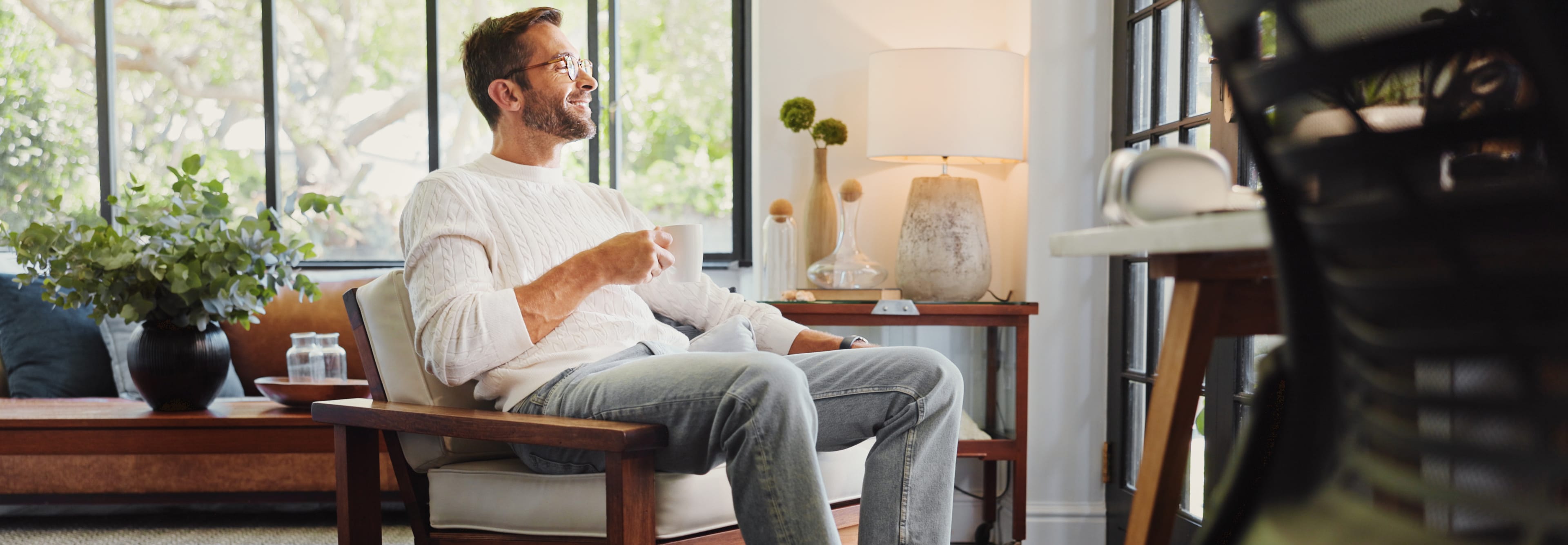 A smiling man with glasses and a white sweater is relaxing in an armchair in a bright living room, holding a mug in his hand.