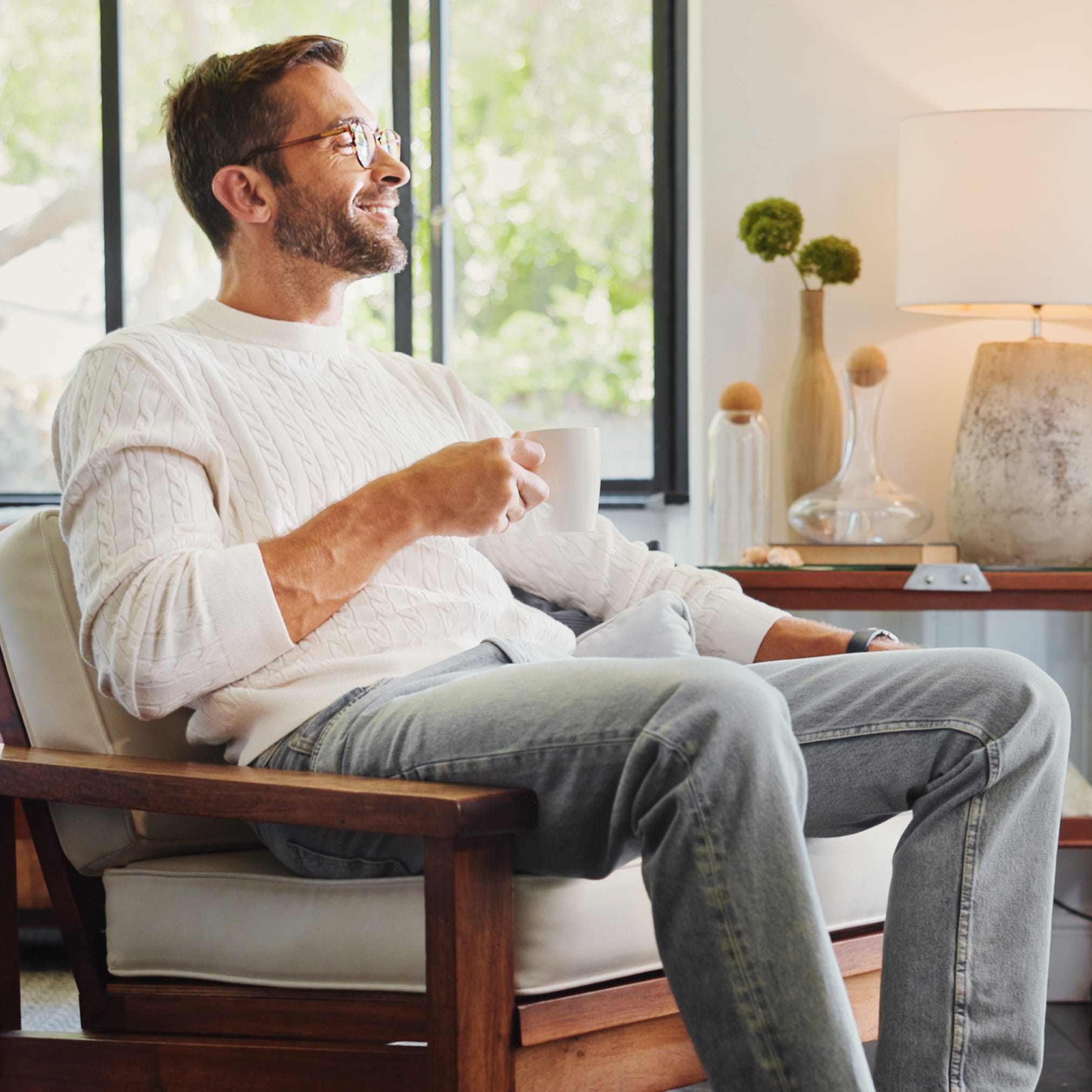 A smiling man with glasses and a white sweater is relaxing in an armchair in a bright living room, holding a mug in his hand.