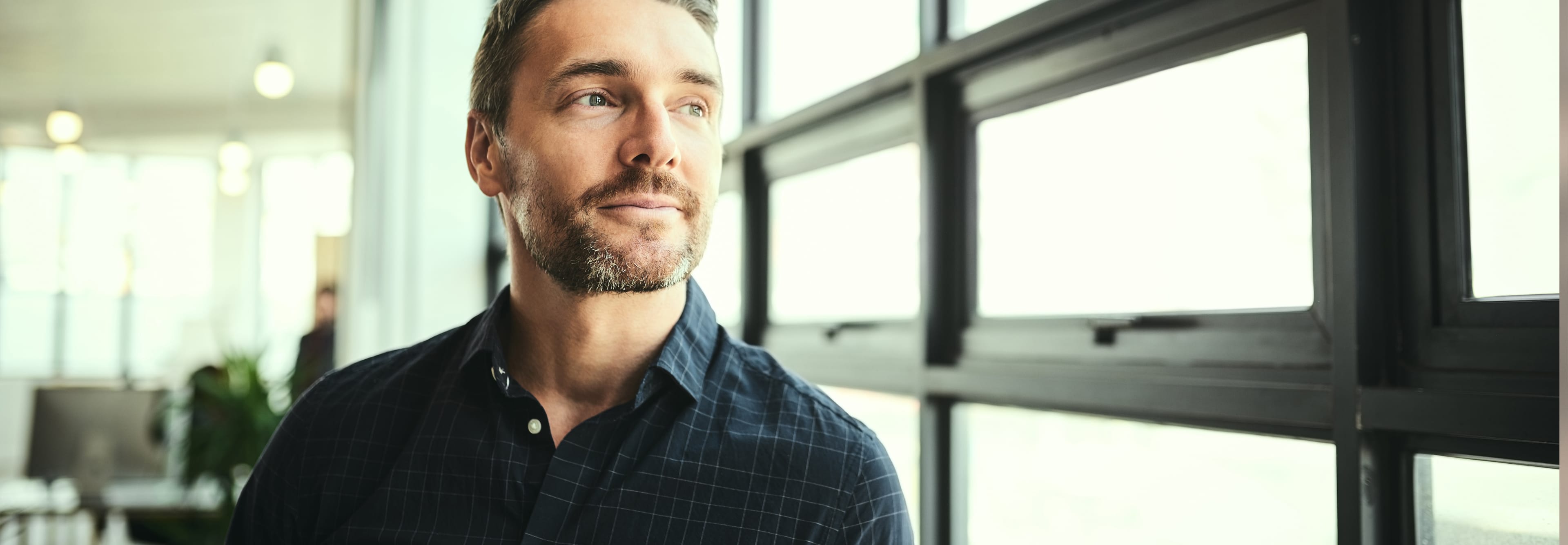 A middle-aged man in a navy blue plaid shirt stands in a bright office and looks with a slight smile through a large window.