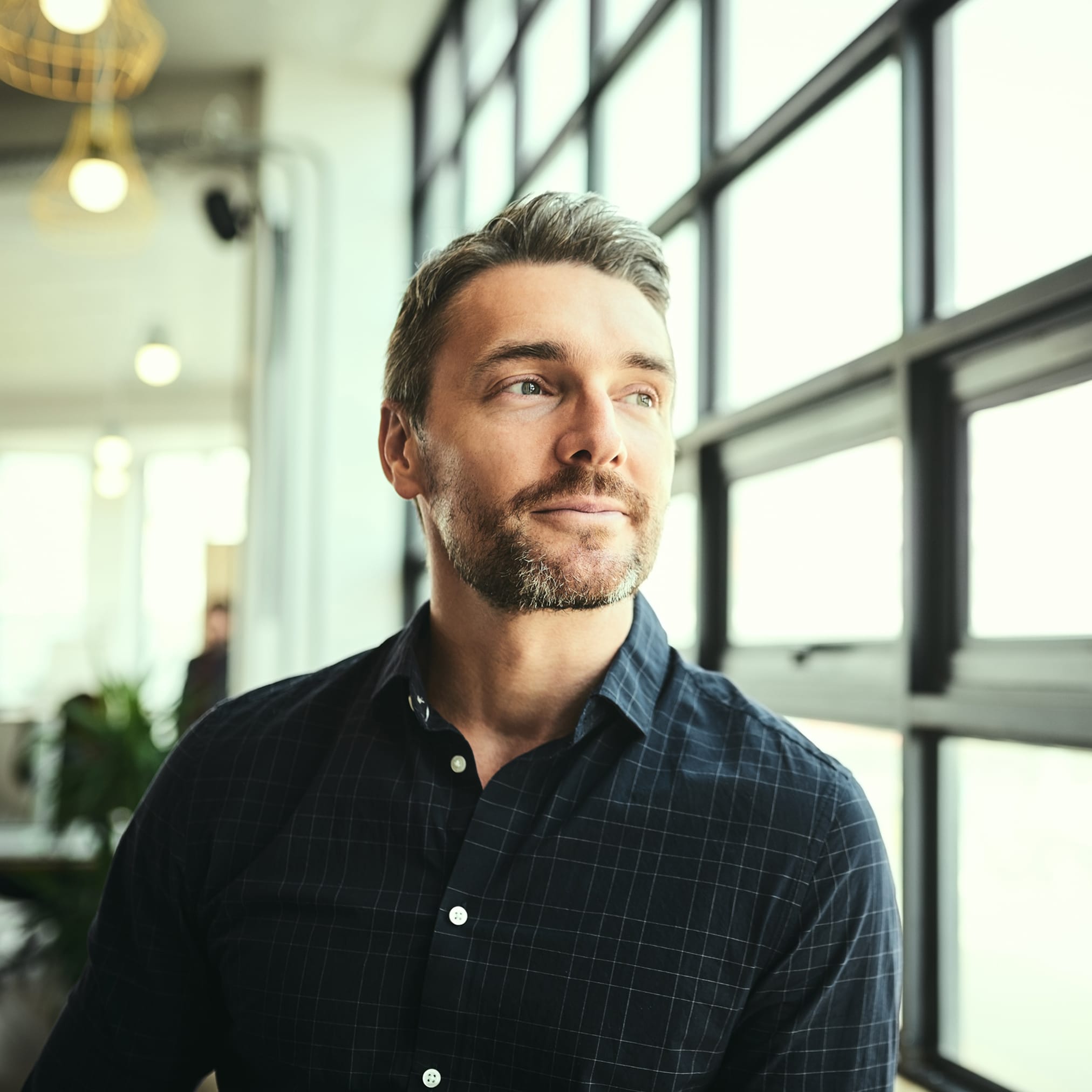A middle-aged man in a navy blue plaid shirt stands in a bright office and looks with a slight smile through a large window.