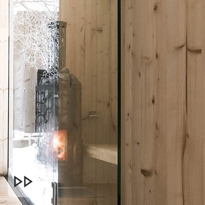 The interior of a sauna finished in light wood, with a black wood-burning stove and a large window with a view of a snowy landscape.