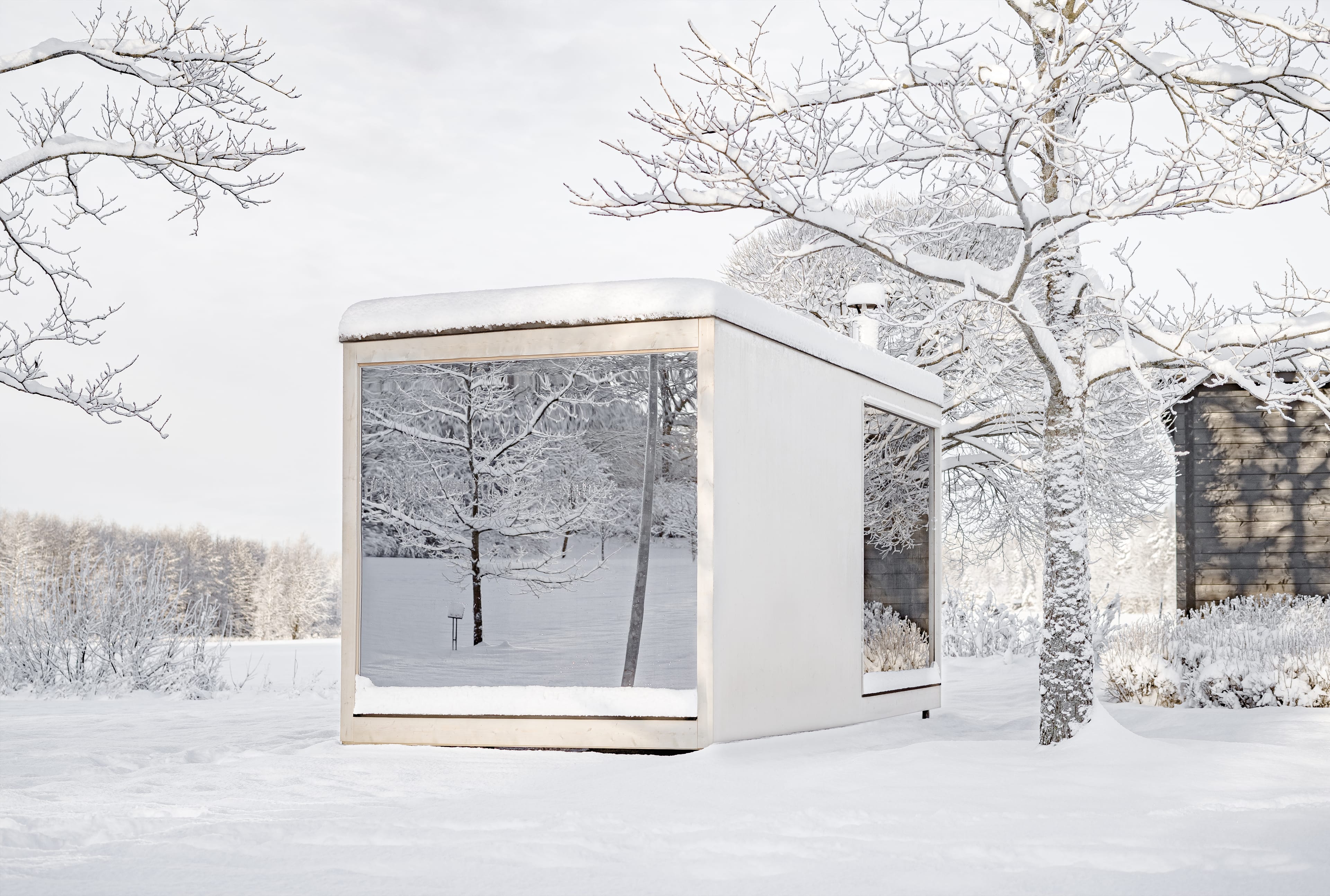 A white, minimalist cabin with panoramic windows stands in a snow-covered garden. The snow-covered trees are reflected in the panes.