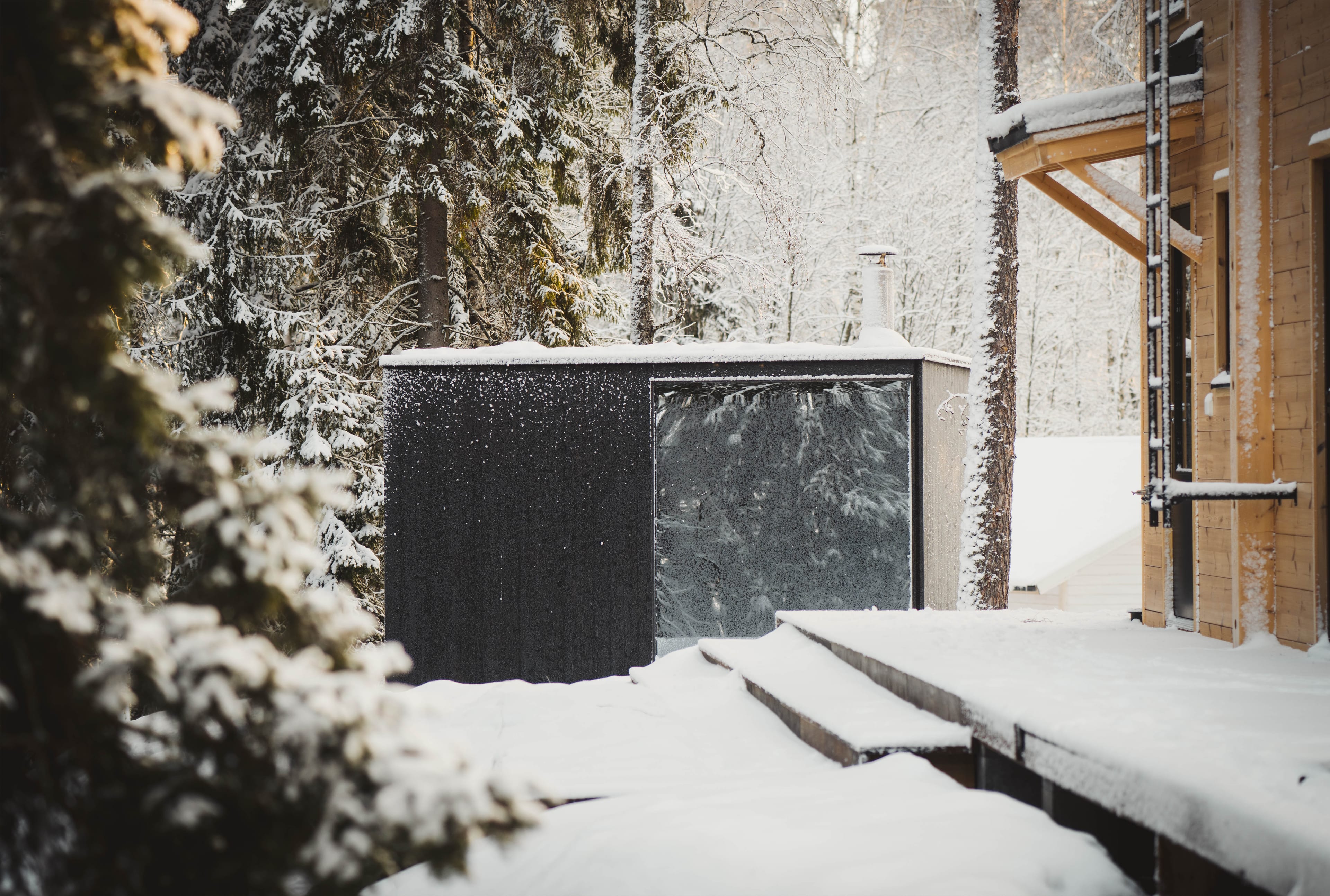 Nowoczesna, zewnętrzna sauna z czarną elewacją i dużym, lustrzanym oknem stoi w ośnieżonym lesie obok drewnianego tarasu.