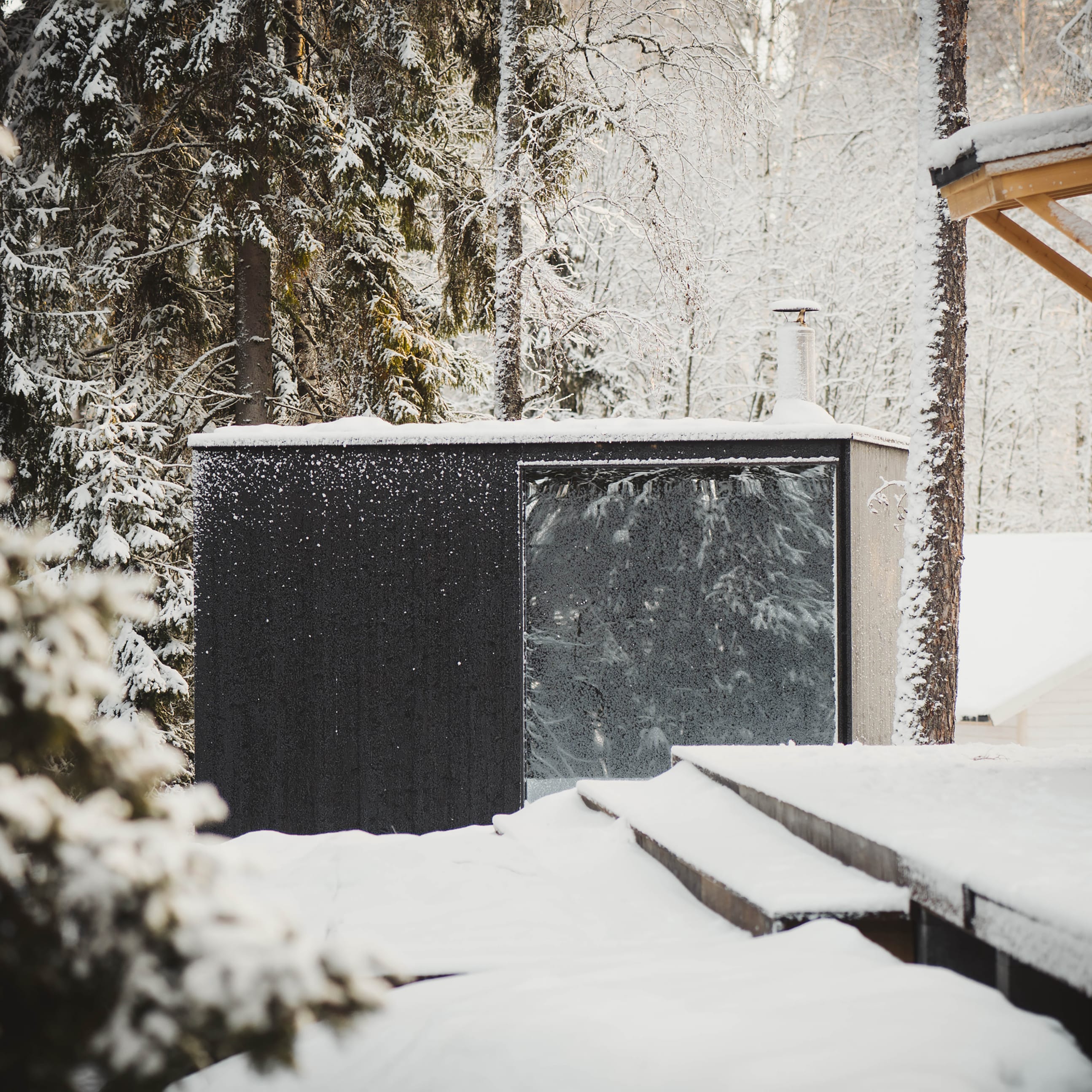 A modern, outdoor sauna with a black facade and a large, mirrored window stands in a snowy forest next to a wooden terrace.