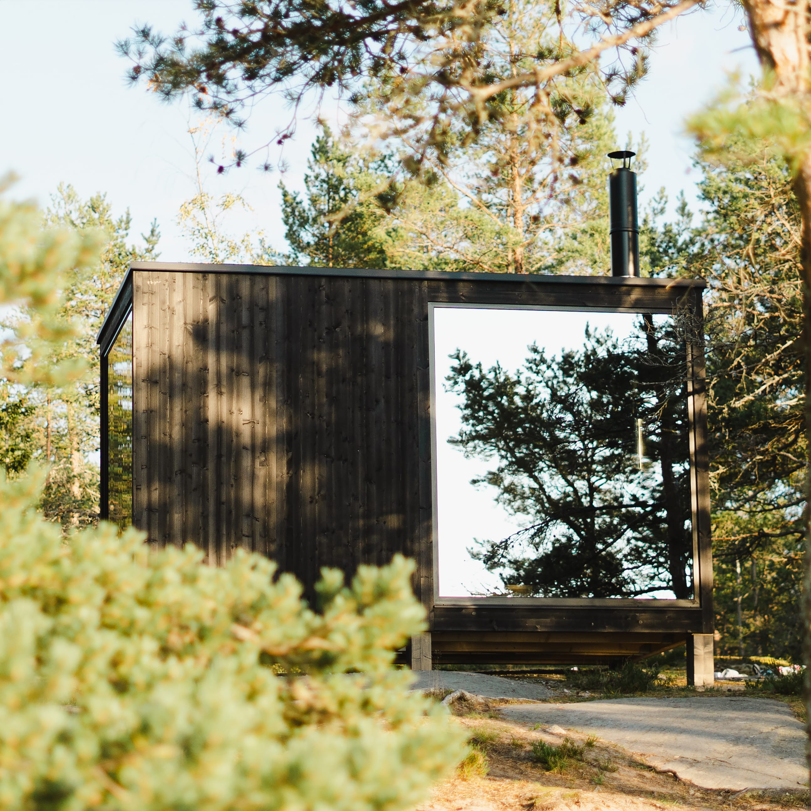 A dark, modern wooden sauna stands in a pine forest. It has a large mirror window reflecting the trees and a chimney on the roof.