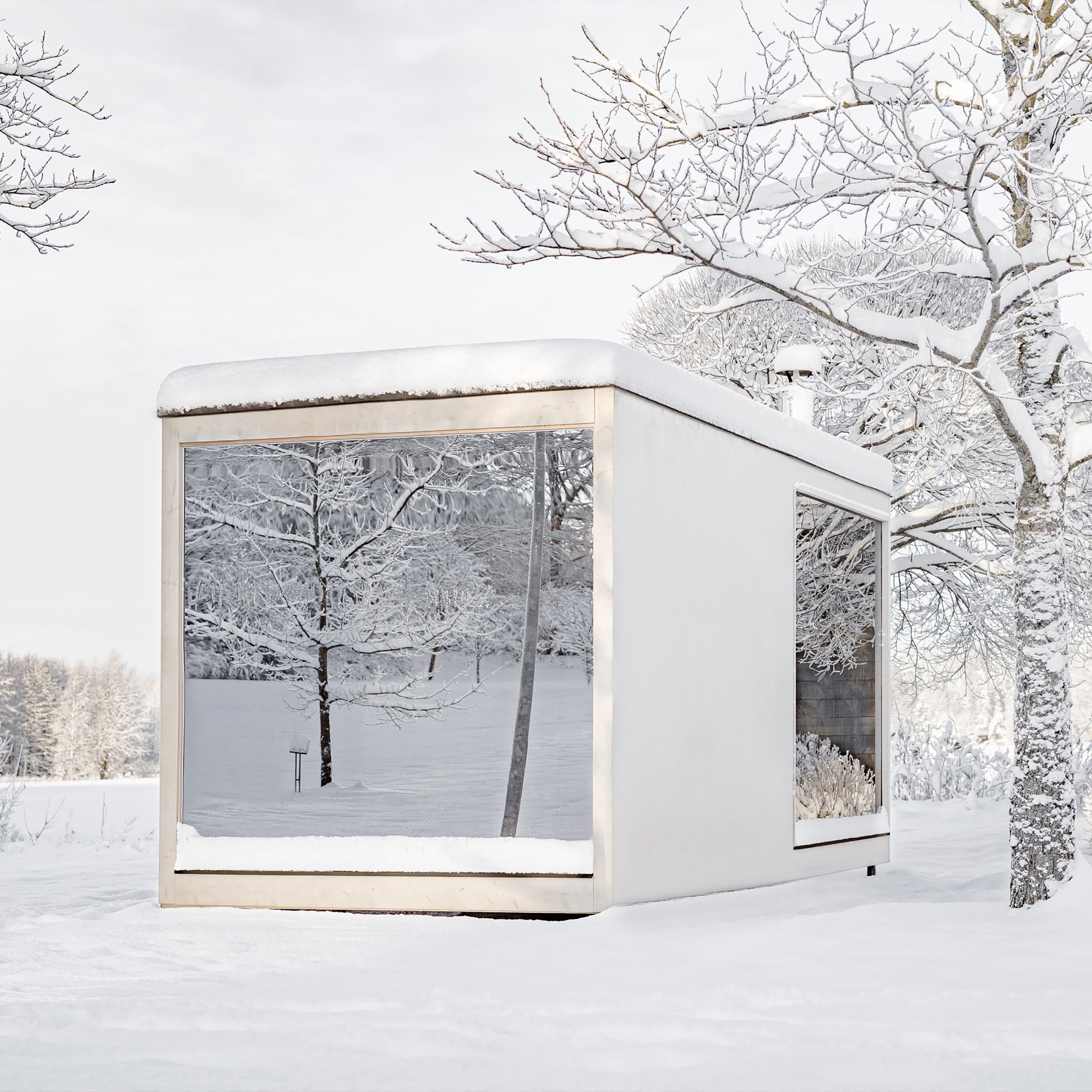 A white, minimalist cabin with panoramic windows stands in a snow-covered garden. The snow-covered trees are reflected in the panes.