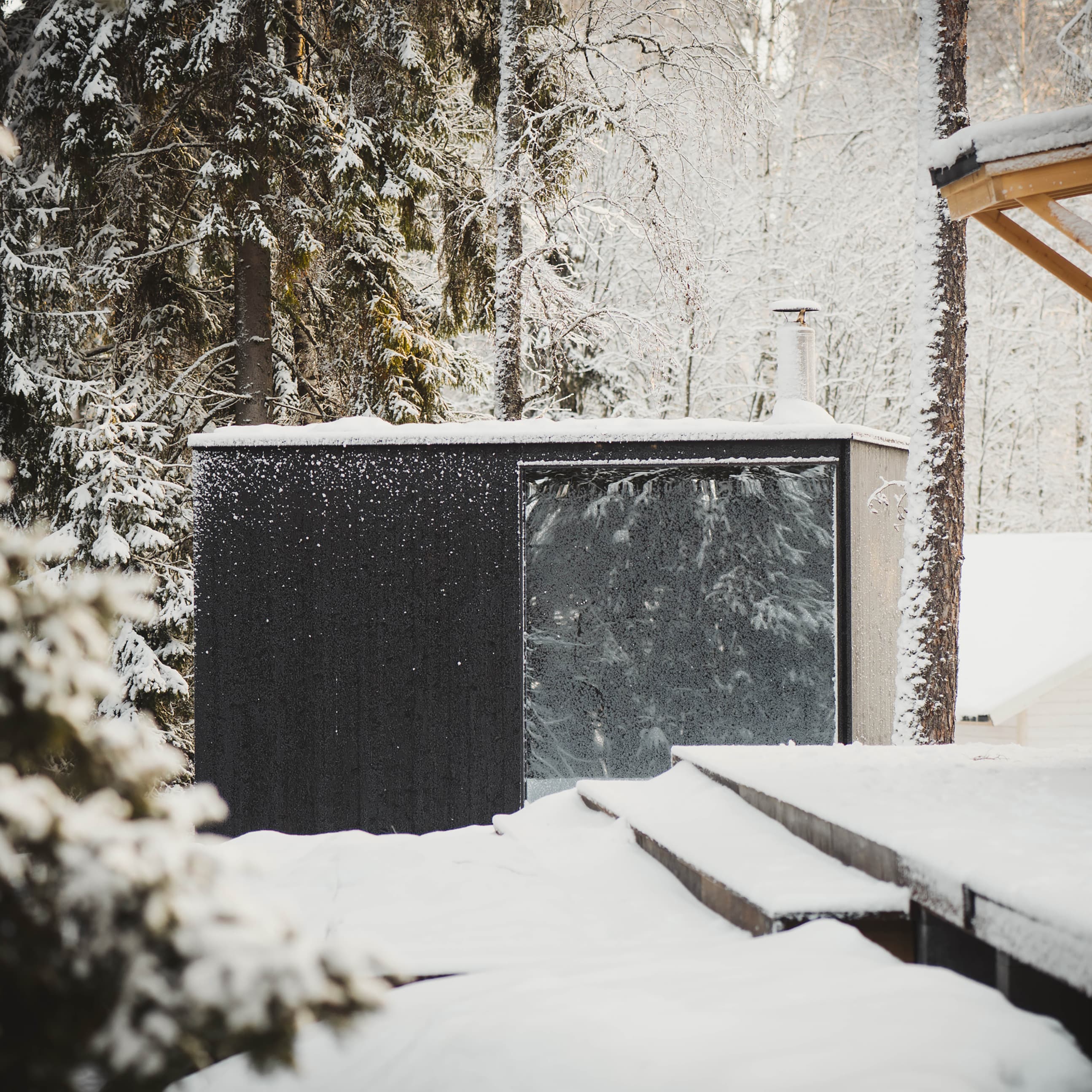 A modern, outdoor sauna with a black facade and a large, mirrored window stands in a snowy forest next to a wooden terrace.