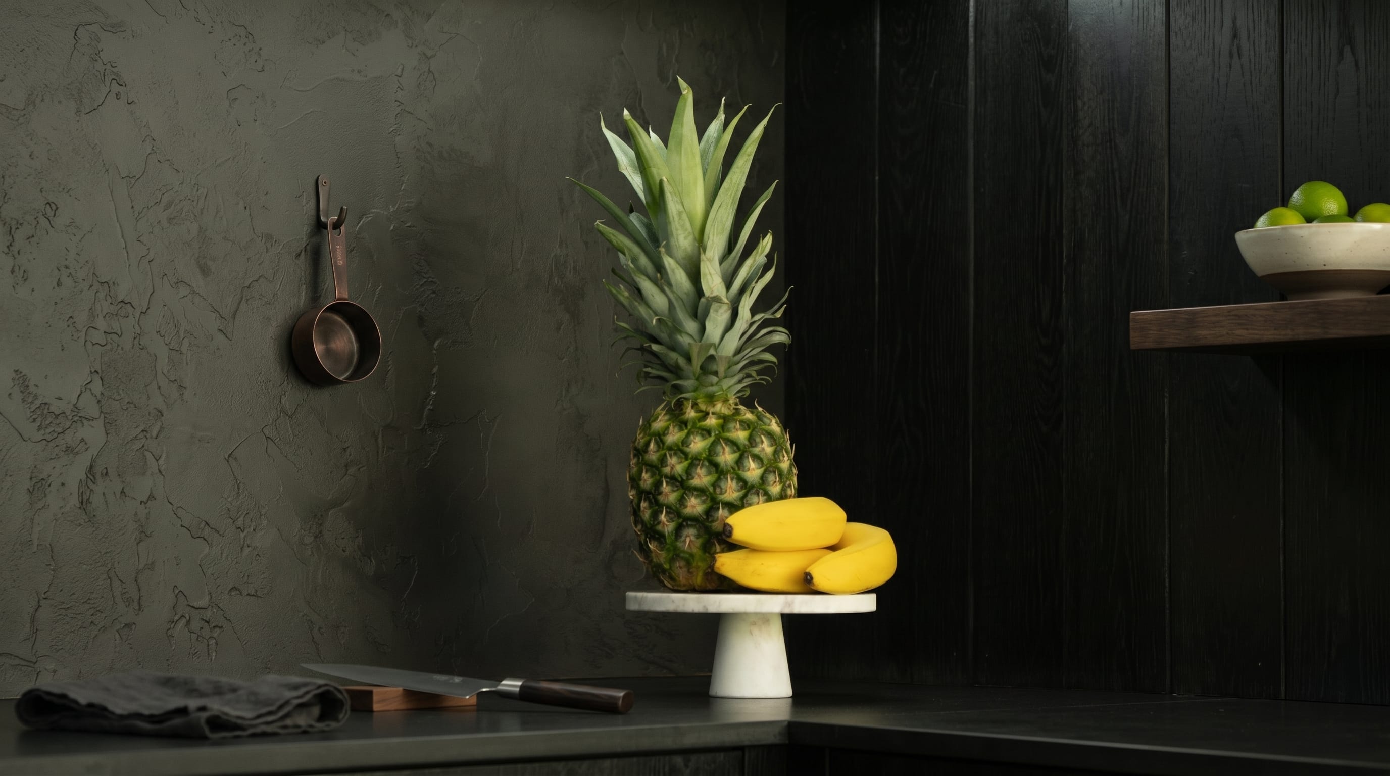 A pineapple and bananas on a stand in a dark Hombee kitchen.