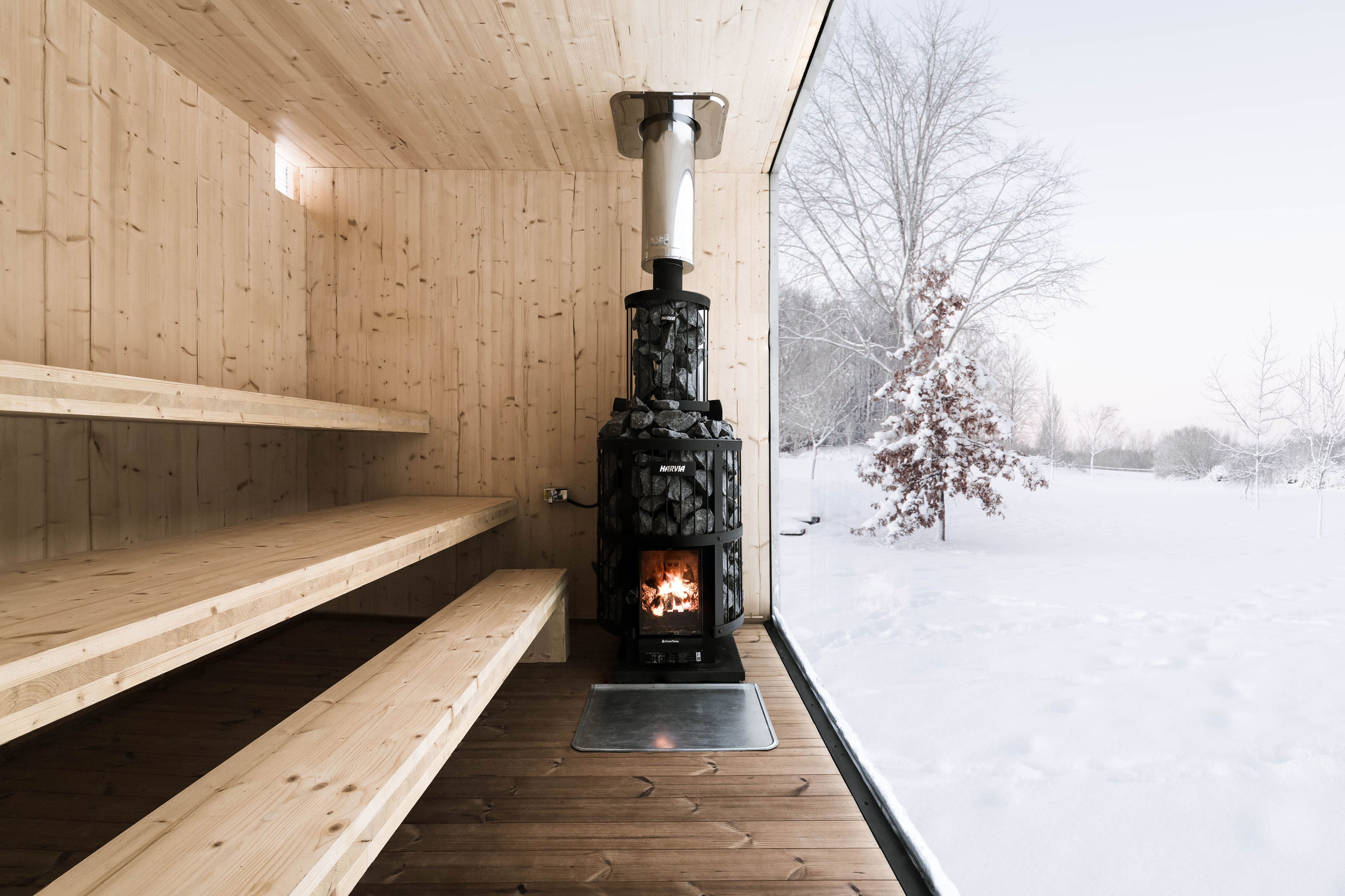 The interior of a sauna finished in light wood, with a black wood-burning stove and a large window with a view of a snowy landscape.