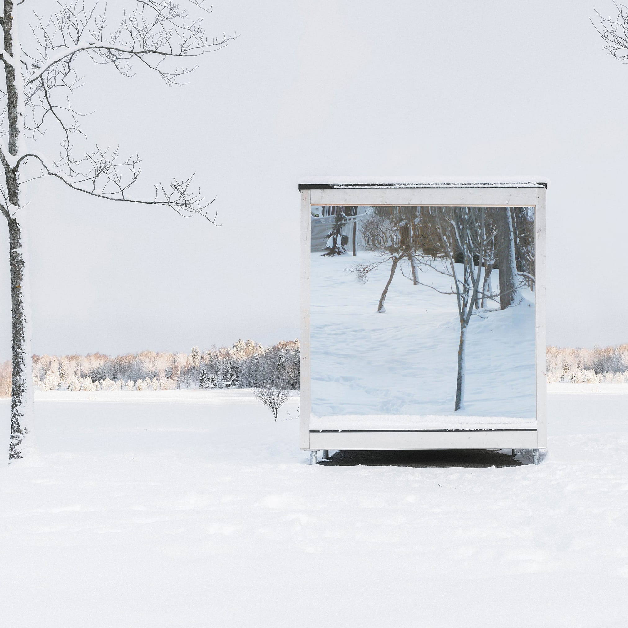 A white, minimalist cabin with panoramic windows stands in a snow-covered garden. The snow-covered trees are reflected in the panes.