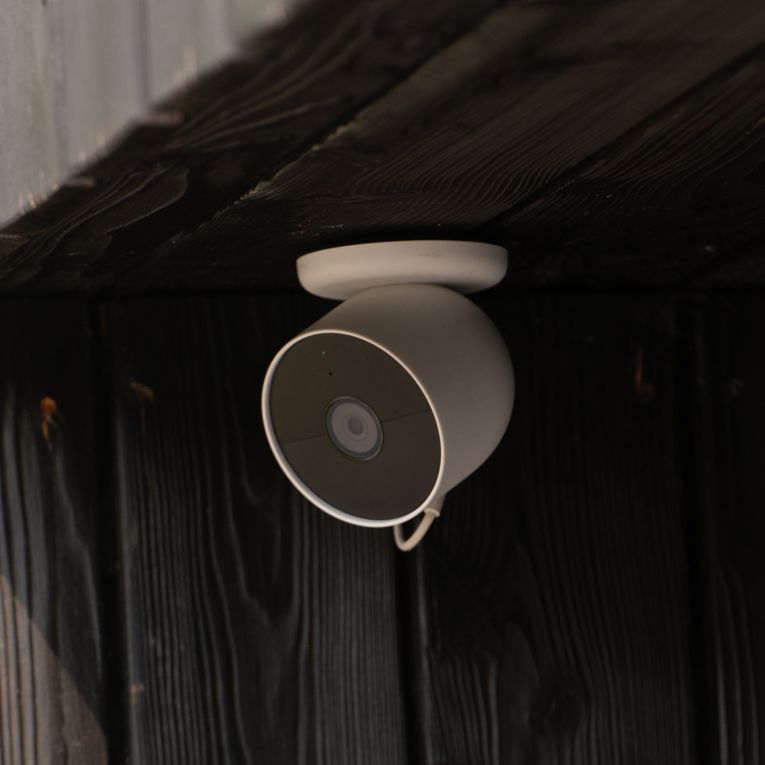 A white, semicircular, discreet camera is mounted in the recess of a house made of dark wood. The camera is in the shade, with the sun's rays falling around it.