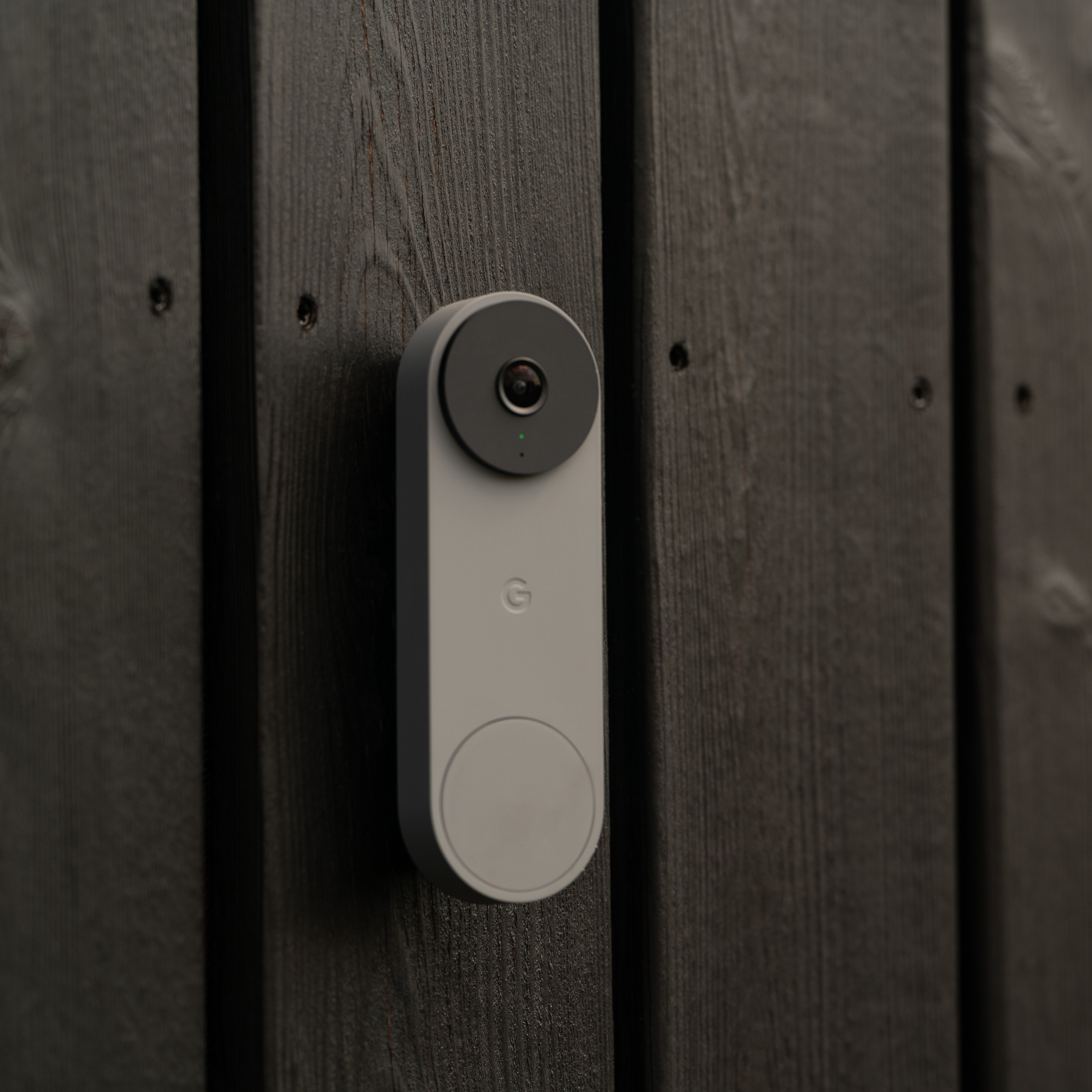 A modern, bright and oblong video doorbell is mounted on a dark, wooden facade. The lens of the video doorbell is clearly visible.