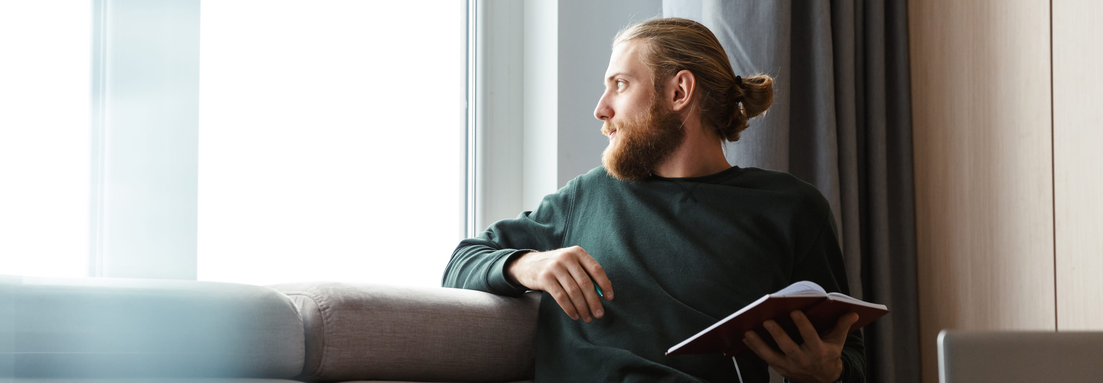 A man with a beard and his hair in a bun sits on the couch. He is holding an open notebook and looking thoughtfully out a large, bright window.