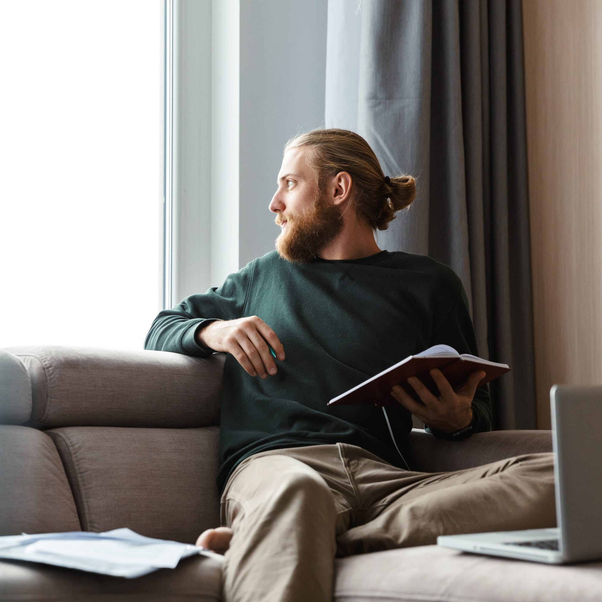 A man with a beard and his hair in a bun sits on the couch. He is holding an open notebook and looking thoughtfully out a large, bright window.