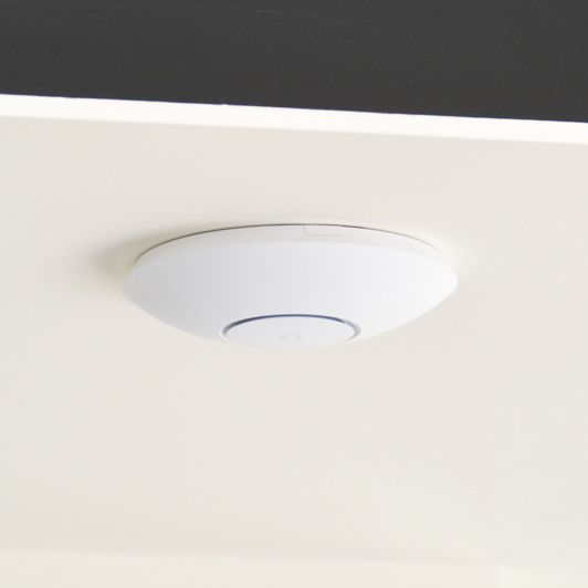 A white, round access point mounted on a light beige ceiling.