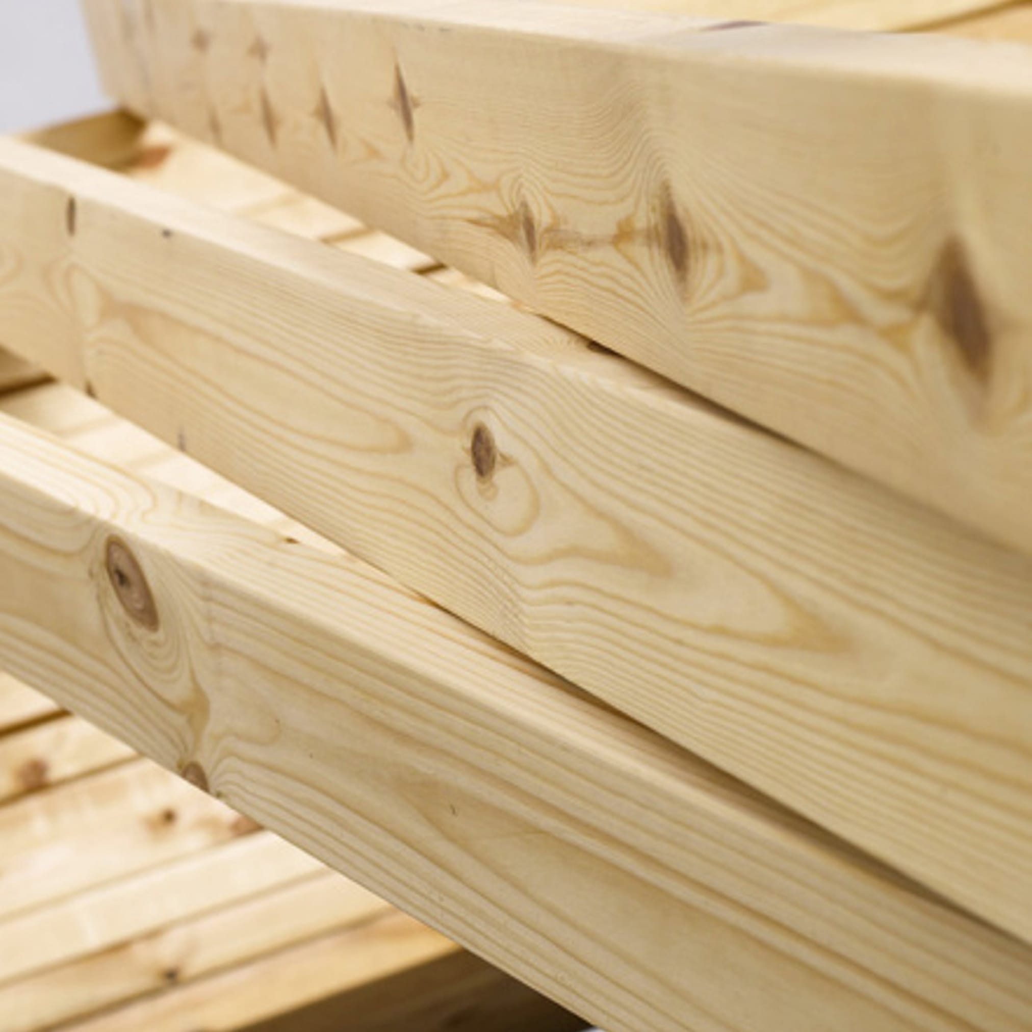 A close-up of a pile of brightly finished structural timber beams, showing the clear grain and knots of the material.